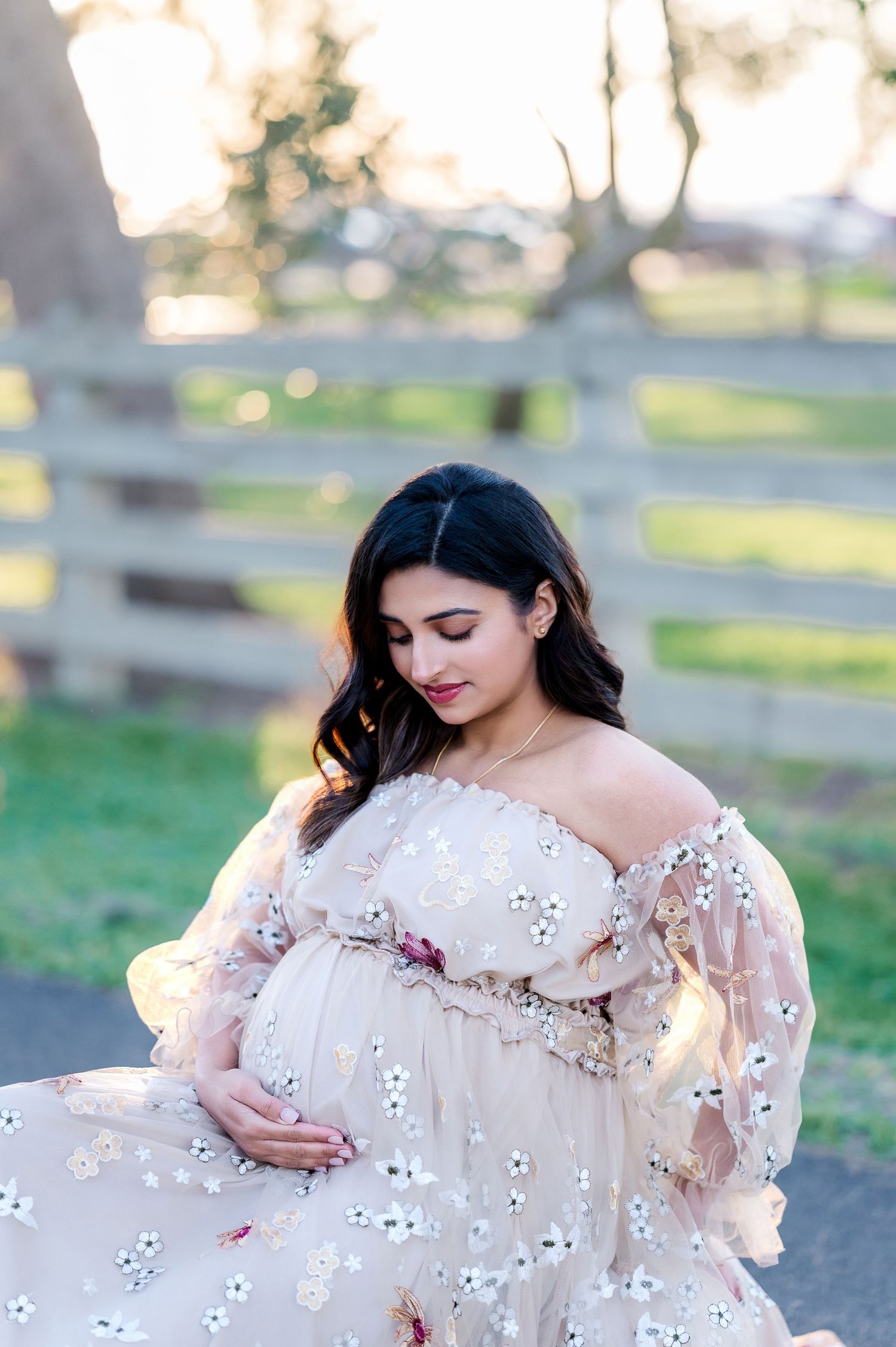 Woman in white eyelet dress poses gracefully against rustic fence during sunset maternity session.