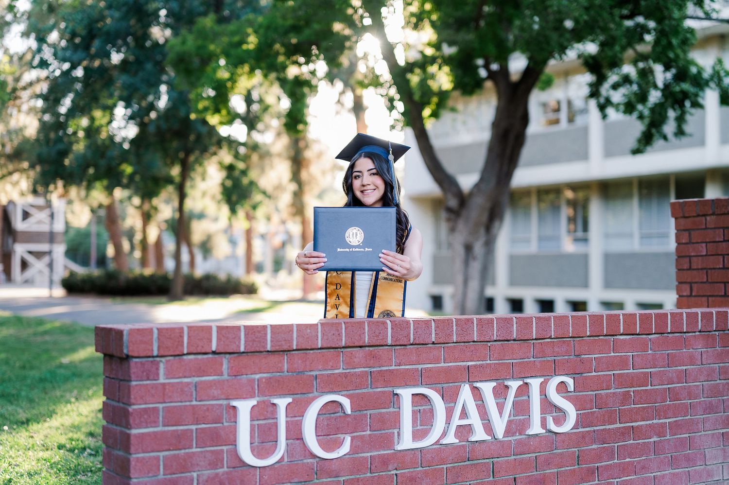 A graduate wearing academic regalia poses on a red brick wall with UC Davis lettering in the background.