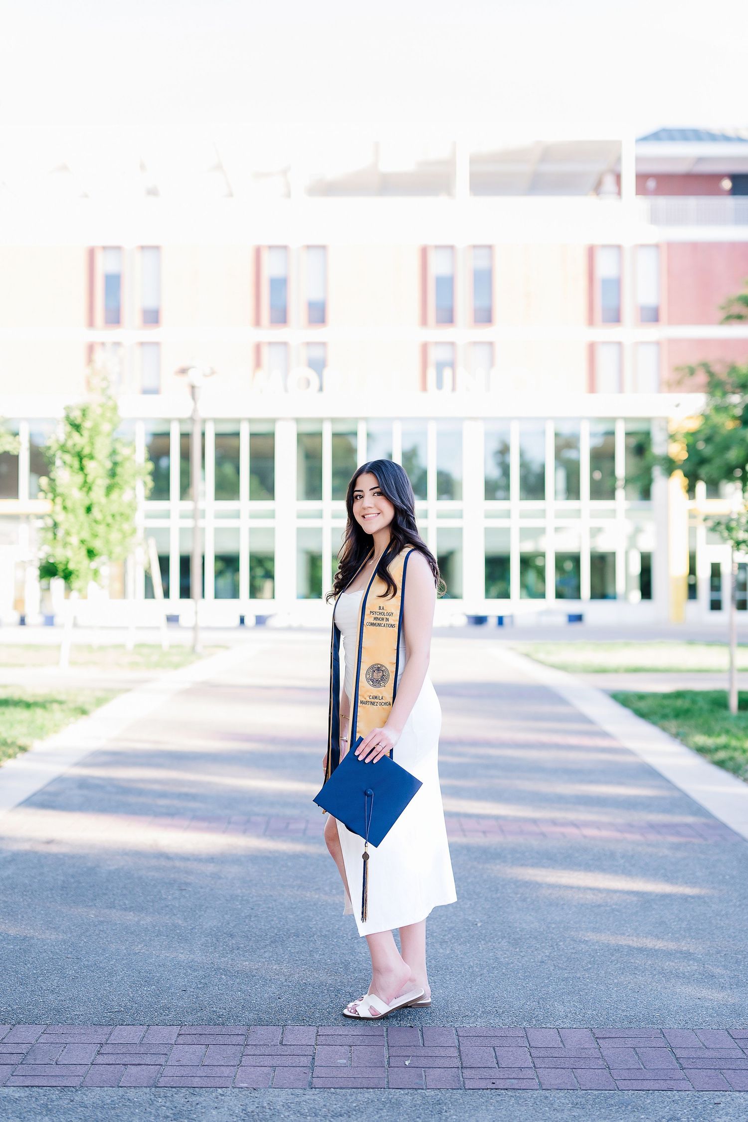 Graduate in white dress carrying diploma and tote bag poses on campus walkway.