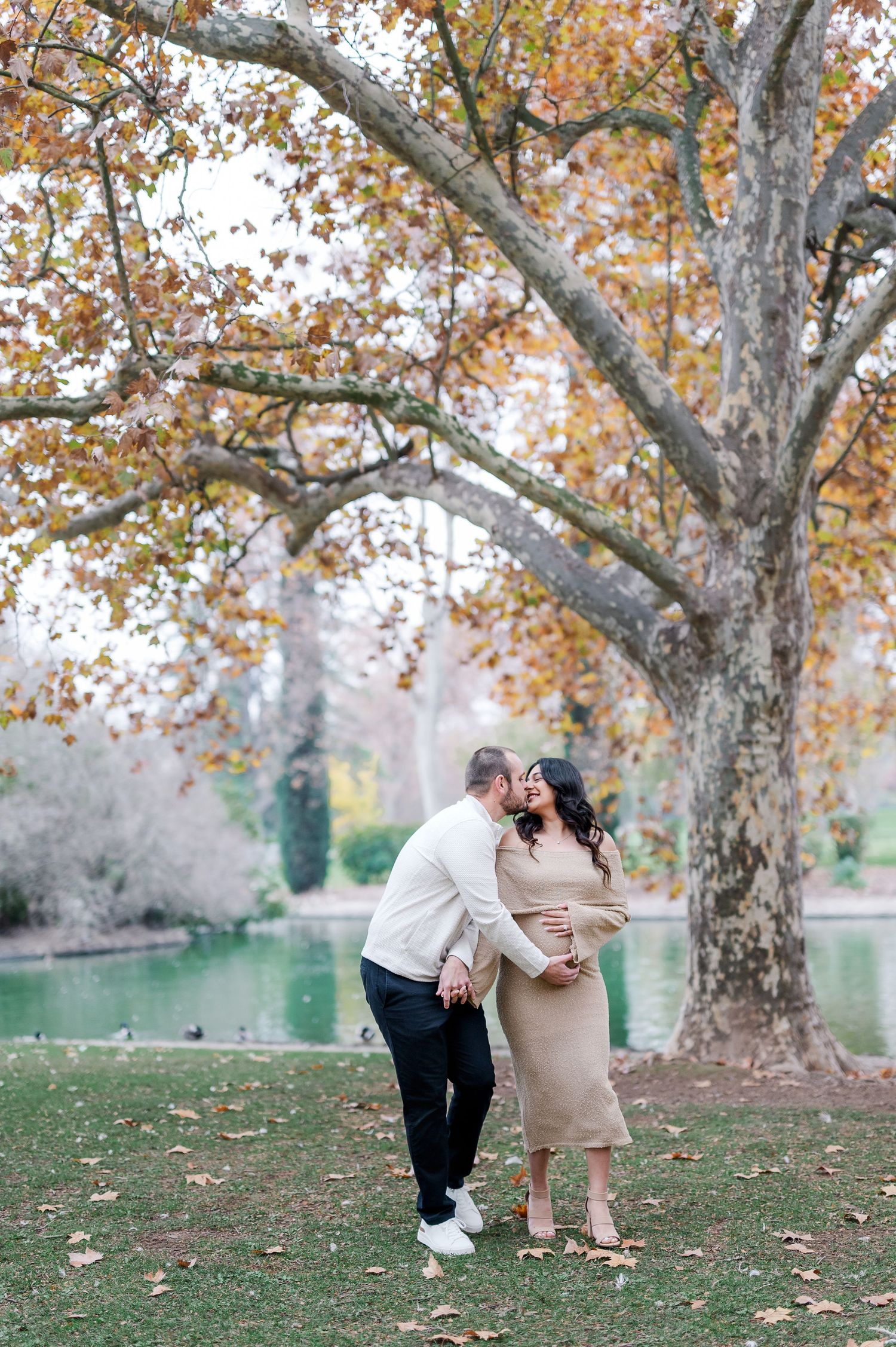 A couple shares a romantic moment under autumn trees with fallen leaves in a park setting.