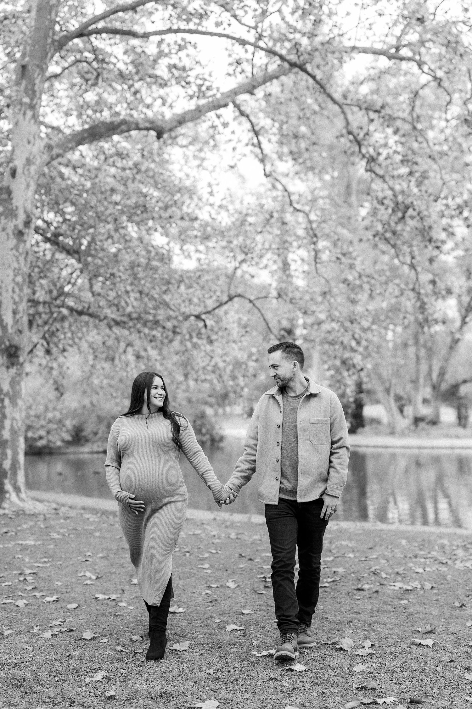 A couple holds hands while walking through a park with autumn leaves scattered on the ground in black and white.