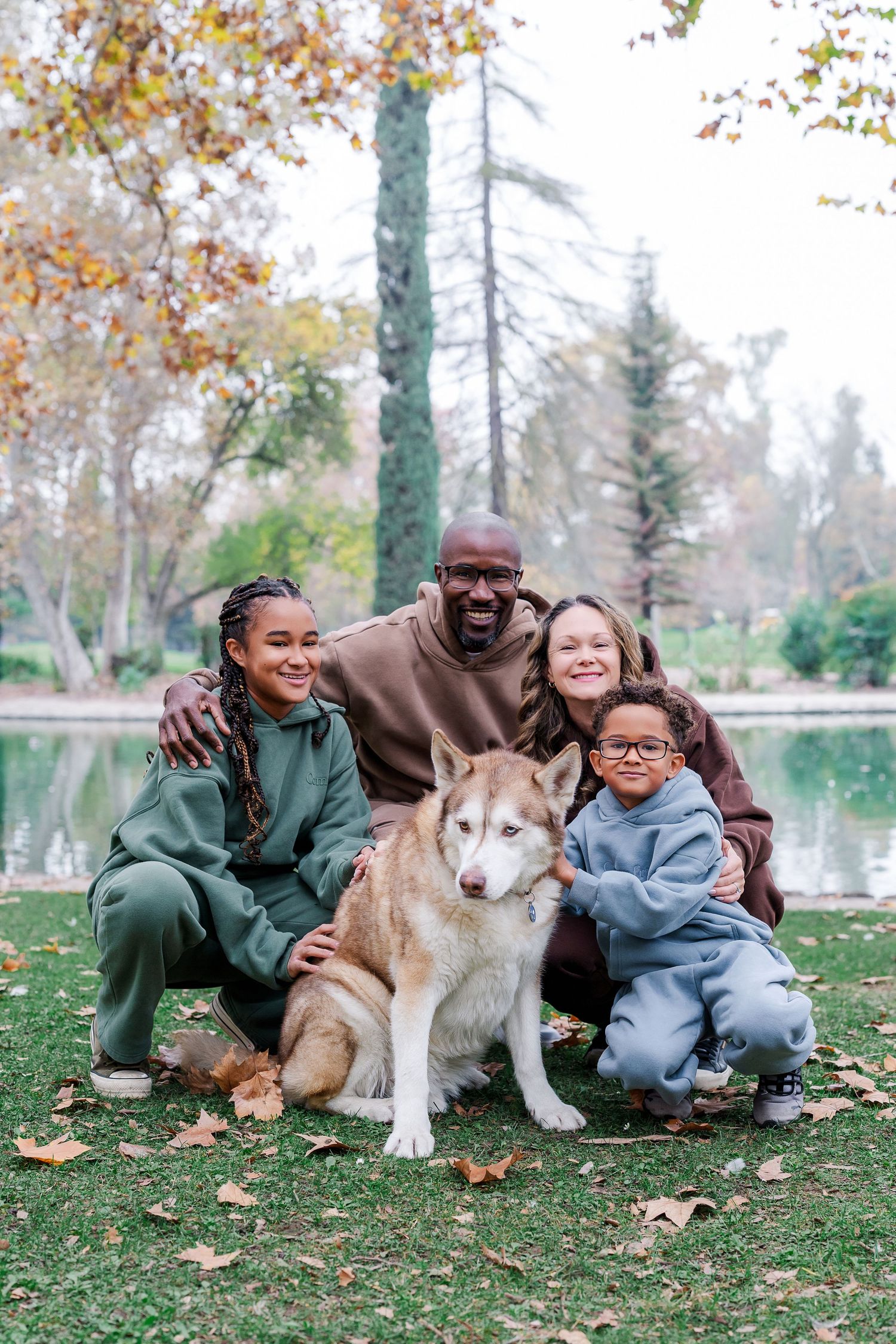 A family sits together with their husky dog on fallen autumn leaves in a park setting.