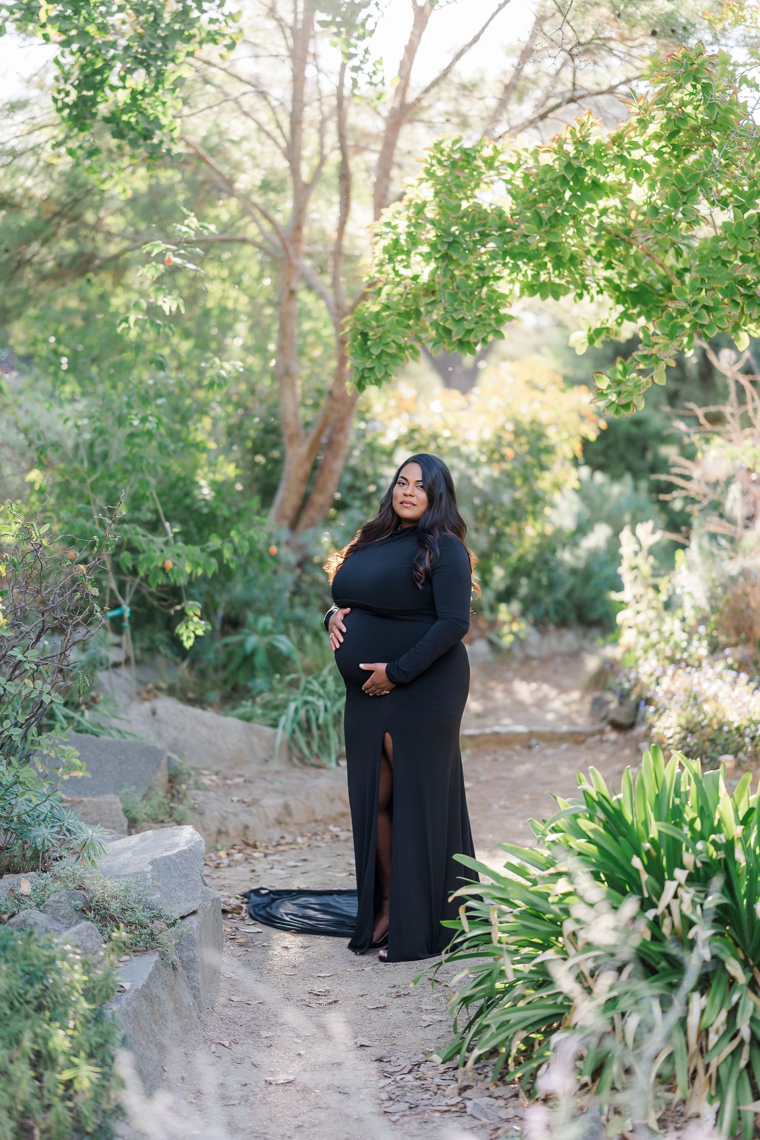 Pregnant woman in elegant black gown cradling baby bump, standing on garden path surrounded by lush green trees.