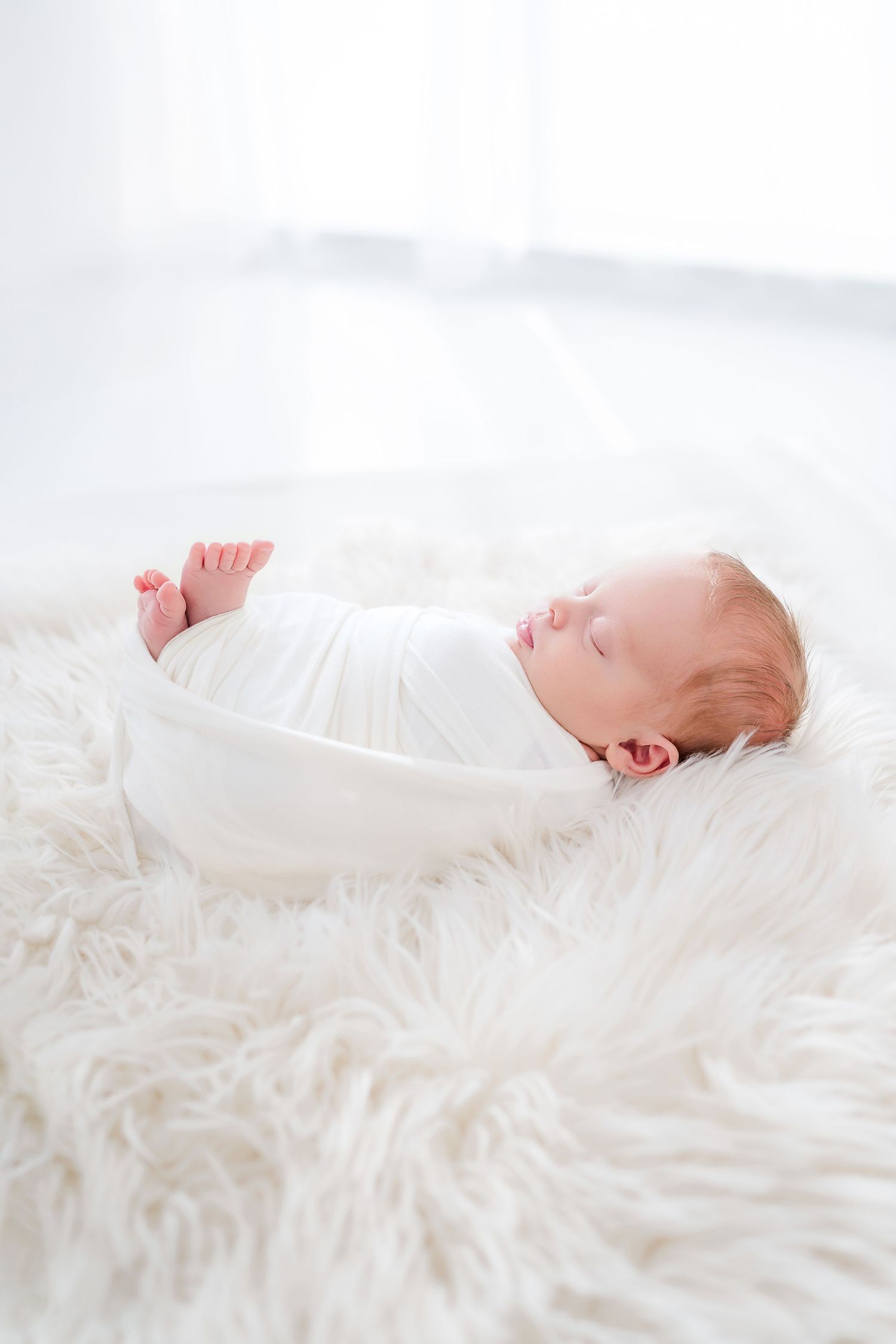 Newborn baby sleeping peacefully on a white fluffy blanket, swaddled in a soft white wrap.