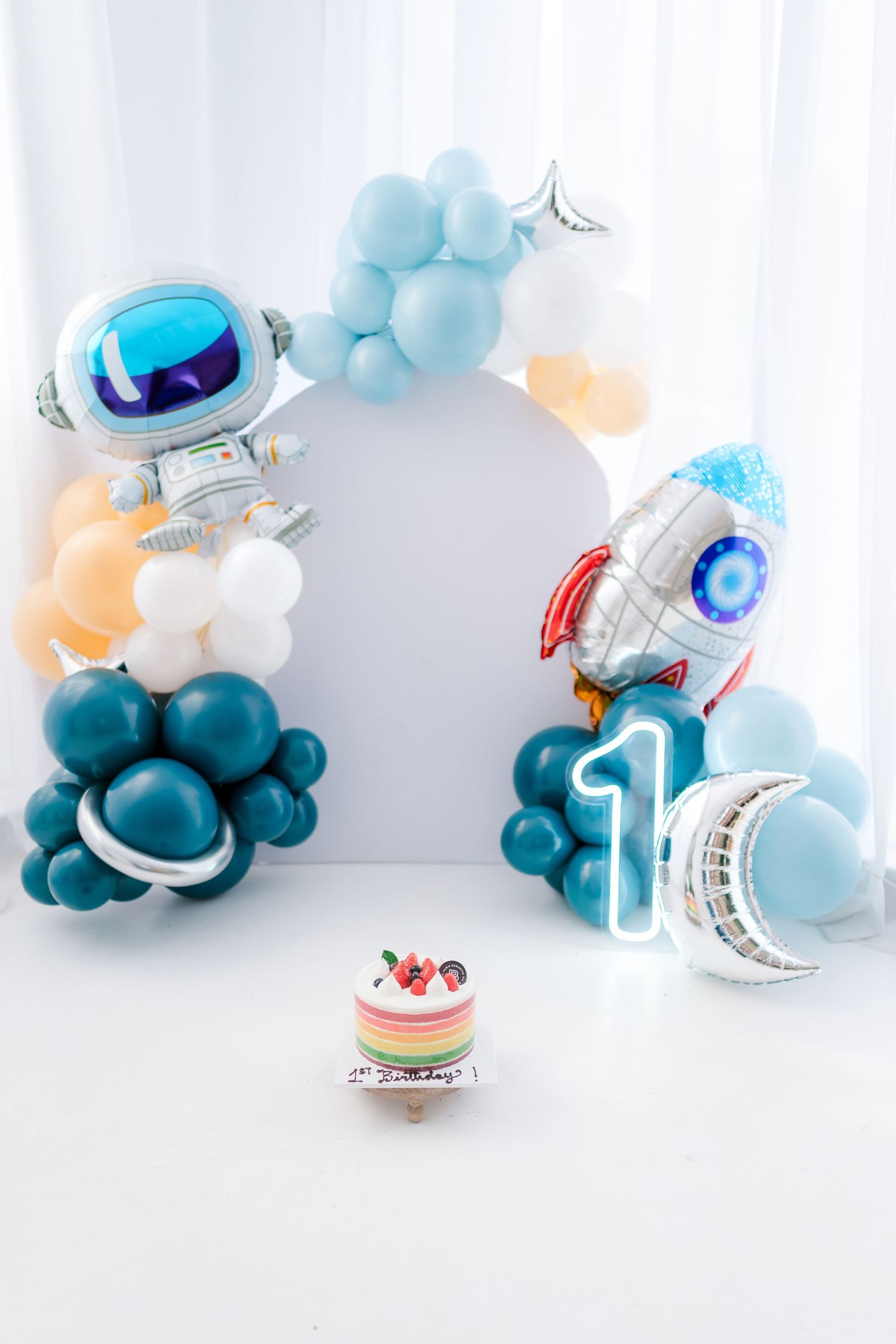 Space-themed party decoration with blue balloon arch, astronaut and rocket balloons, and a small birthday cake centerpiece.