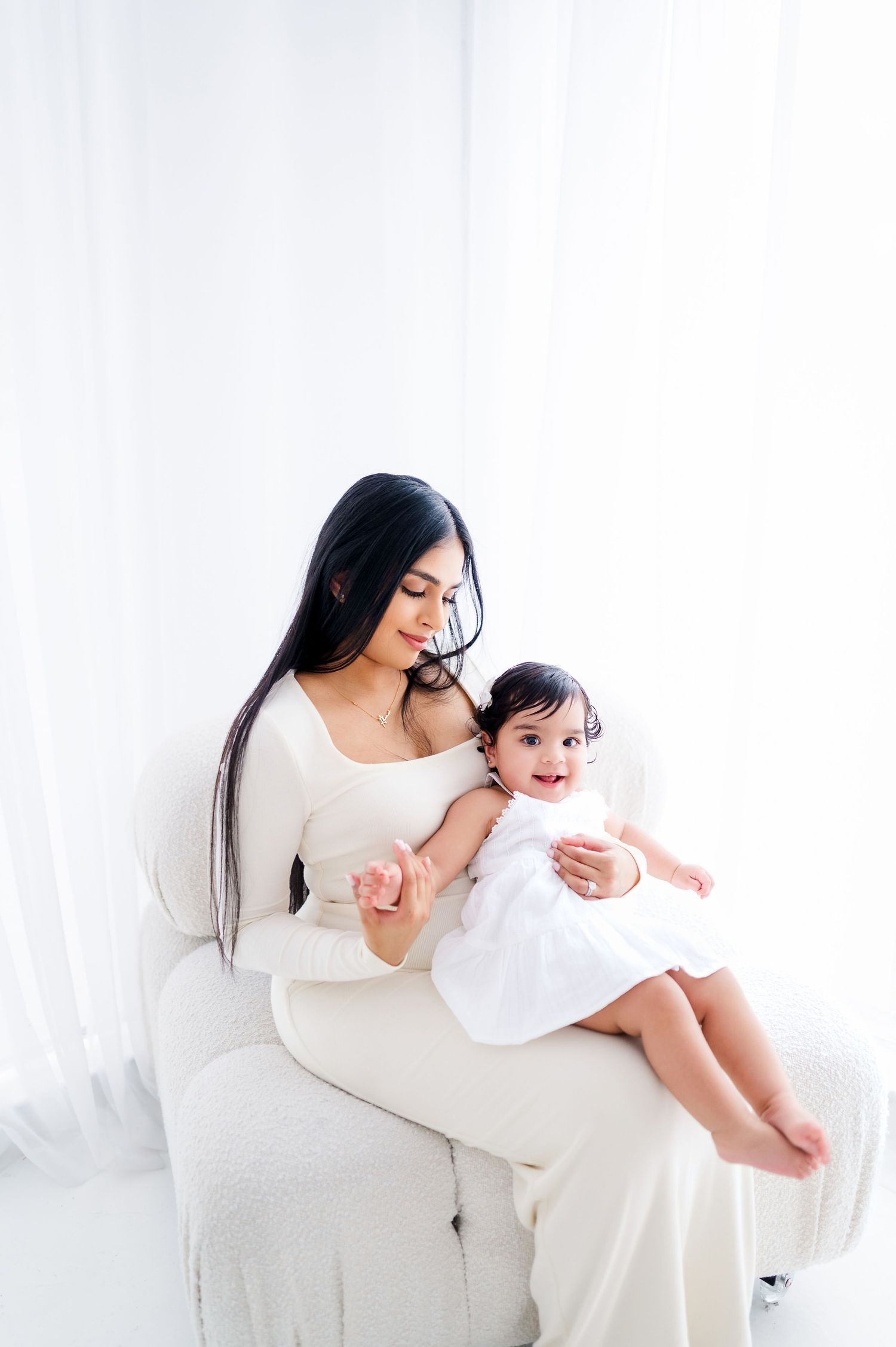 Mother in white dress holding happy baby on white couch during photo session.