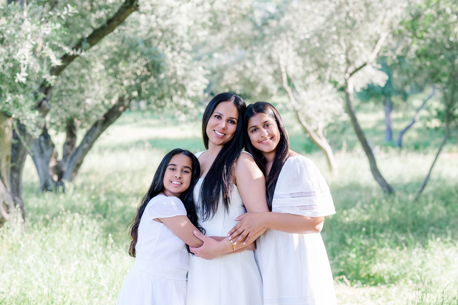 Three people in white outfits embracing in a sun-dappled orchard setting.