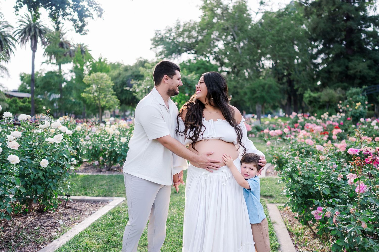 Couple with young child posing in rose garden during maternity photo shoot, woman in white dress cradling baby bump.