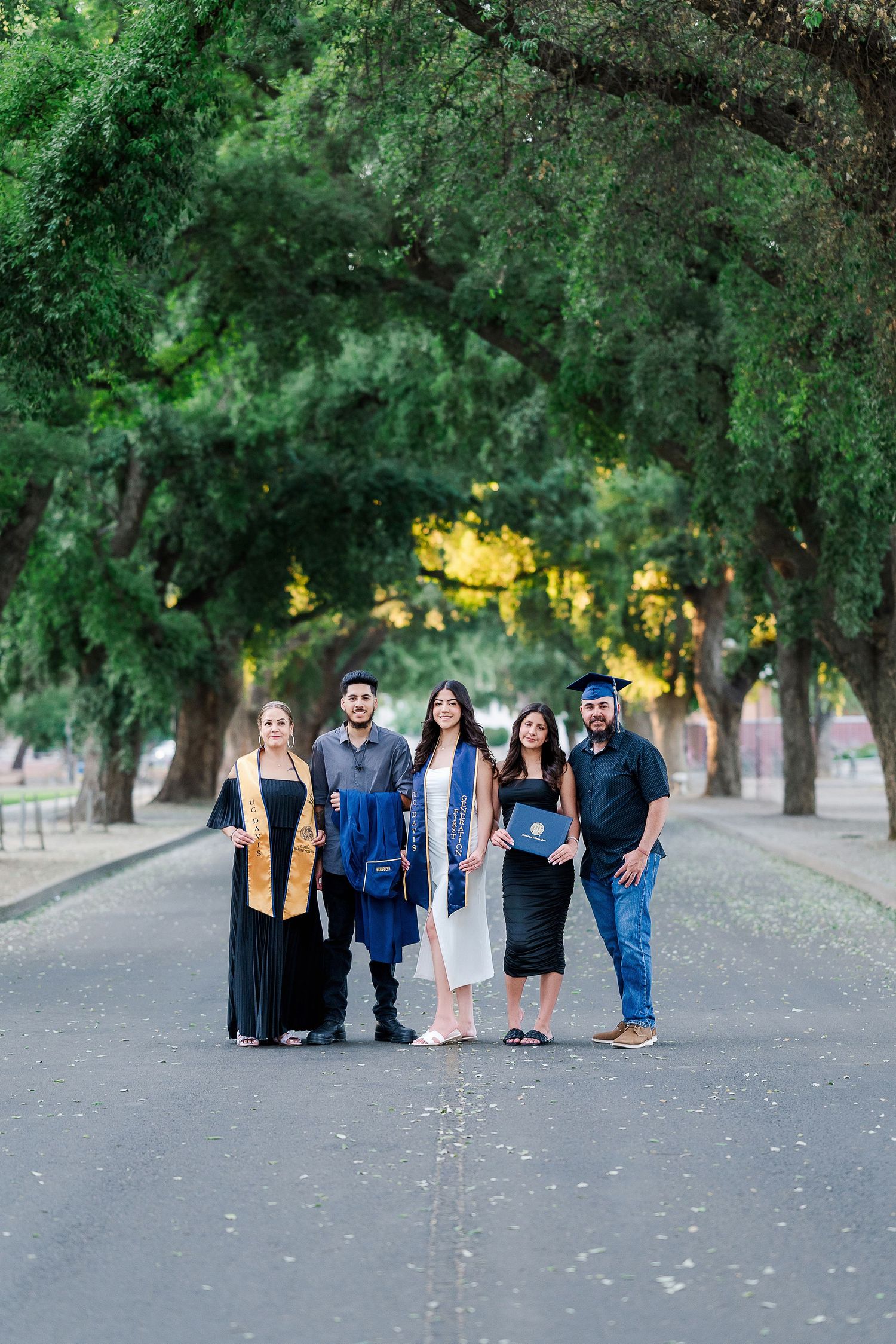 Group of graduates in caps and gowns posing together under trees with string lights at dusk.
