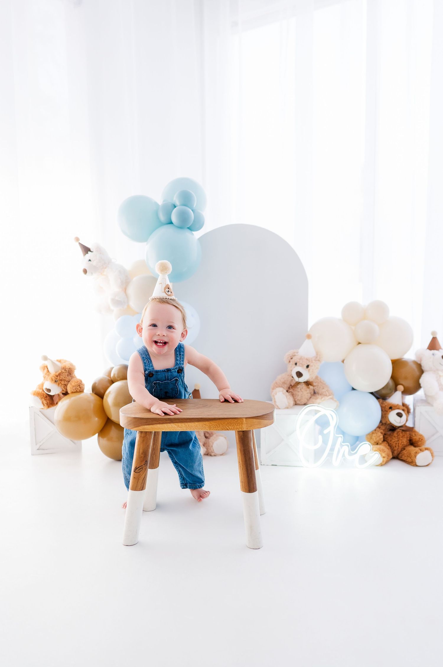 Smiling baby in denim overalls and party hat stands at wooden stool surrounded by balloons and teddy bears at first birthday.