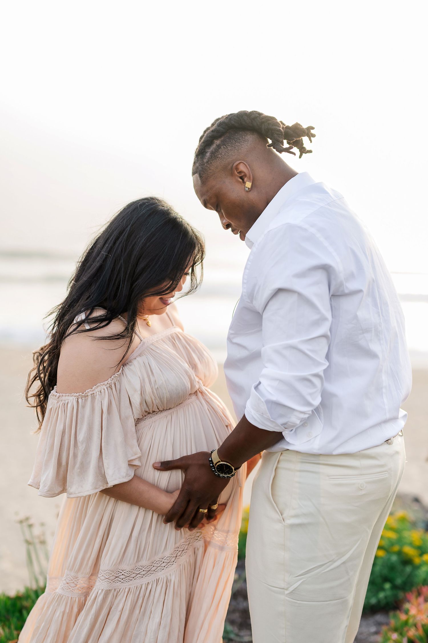 Intimate maternity beach photo with couple in white and beige outfits at sunset.