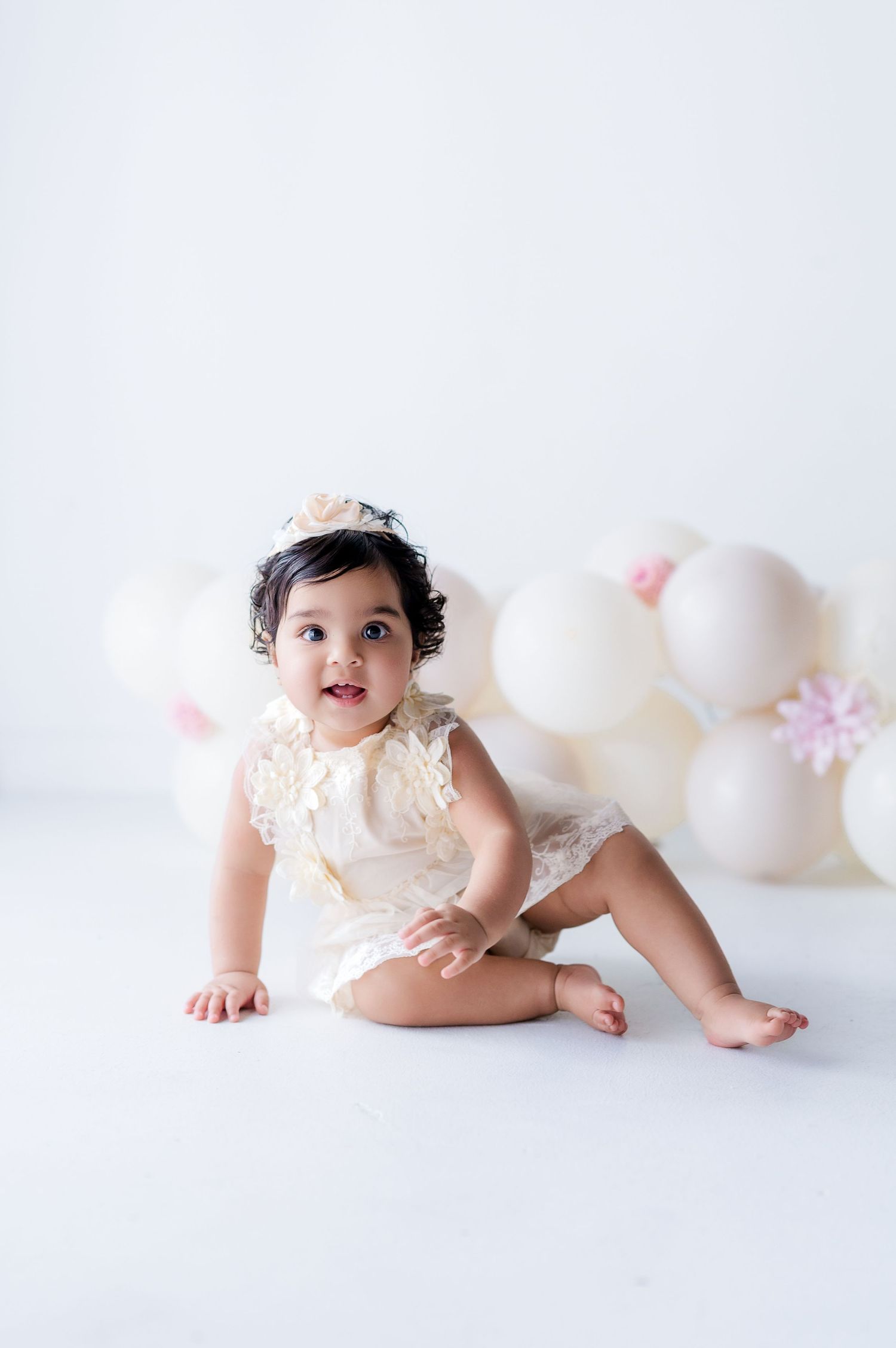 Baby in white lace romper sitting against white background with balloons.