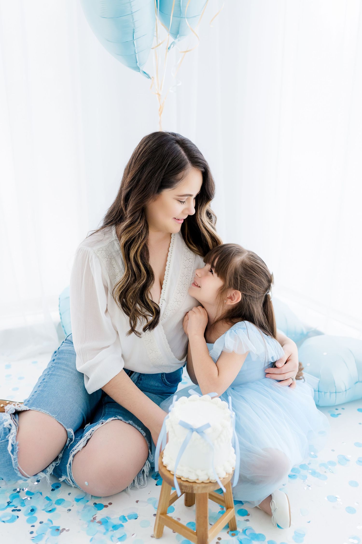 Mother and daughter share a tender moment beside a blue birthday cake, surrounded by blue balloons and confetti.