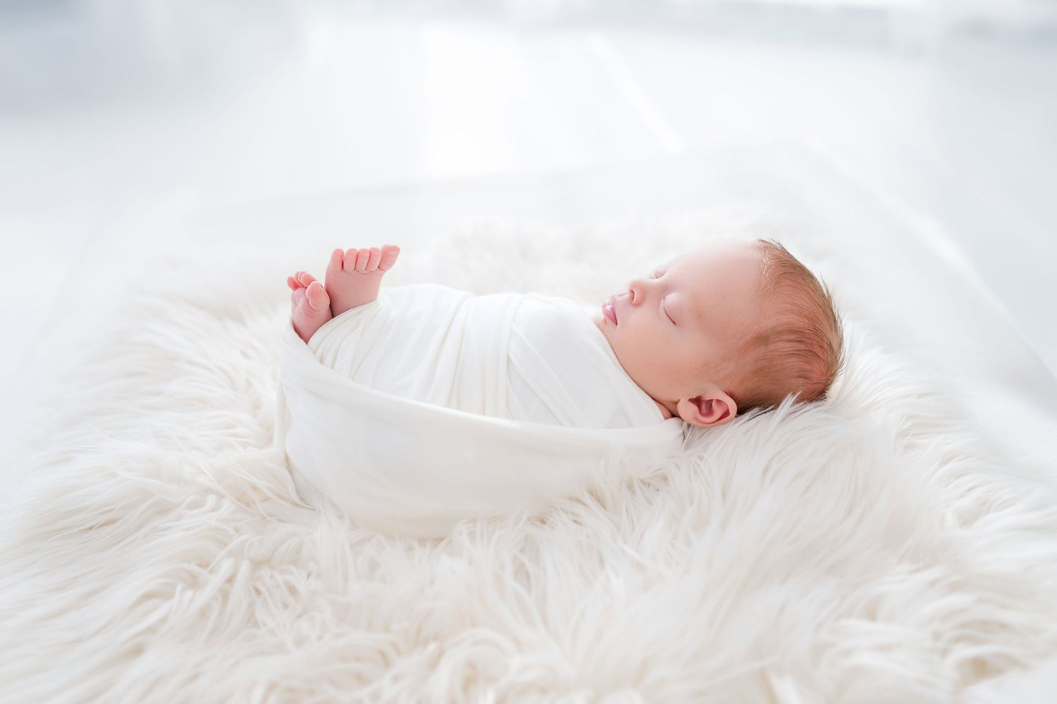 Peaceful newborn baby swaddled in white blanket sleeping on fluffy white fur rug in bright airy studio. Sacramento Newborn Photographer