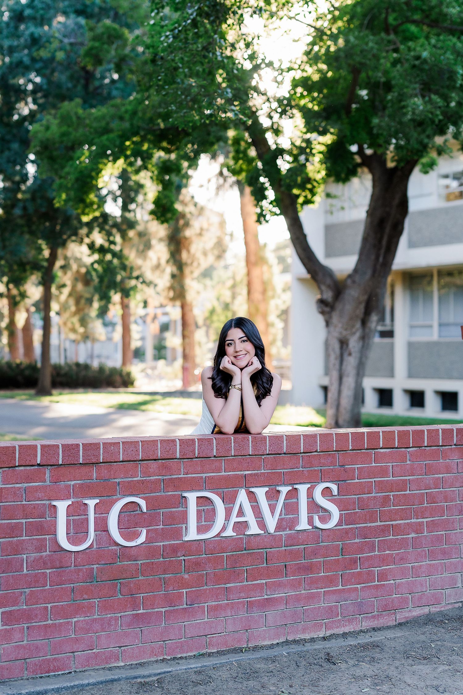 Graduate poses creatively behind red brick UC Davis entrance sign.