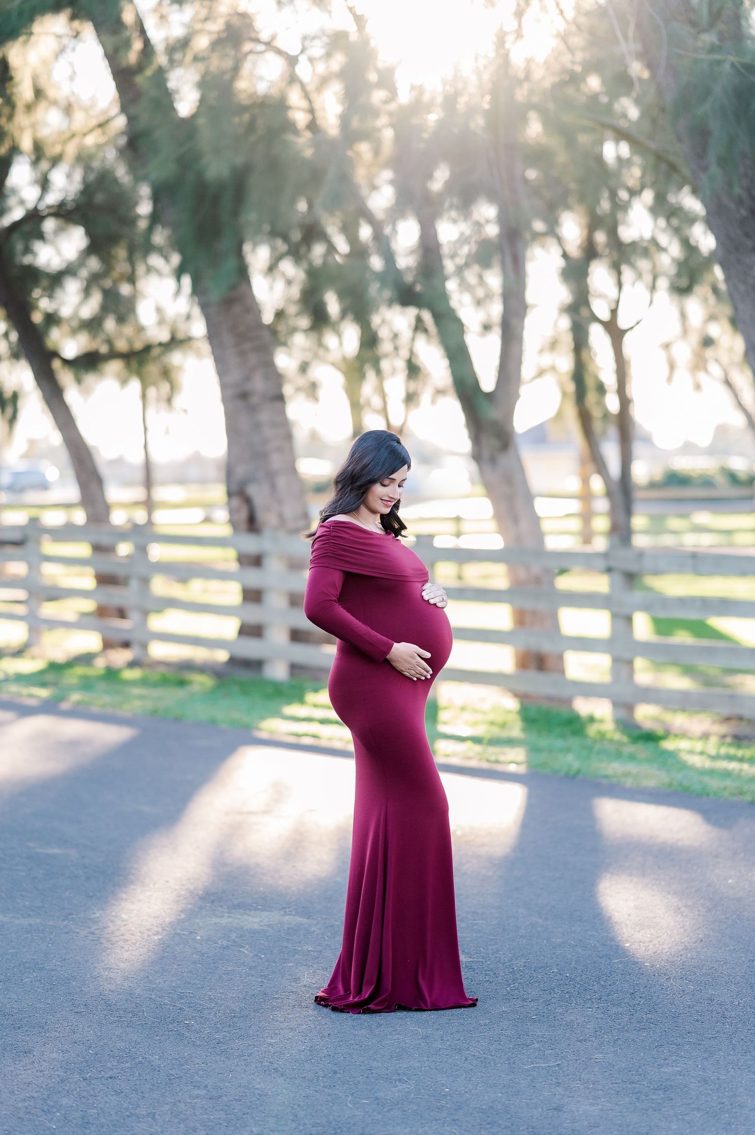 Maternity photography session showing woman in burgundy dress posing in sunlit park setting.