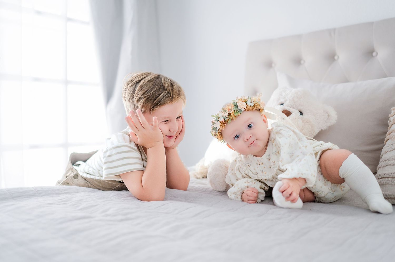 A smiling toddler boy lies on a bed facing a baby girl in a floral crown and white dress, with a stuffed bear nearby.
