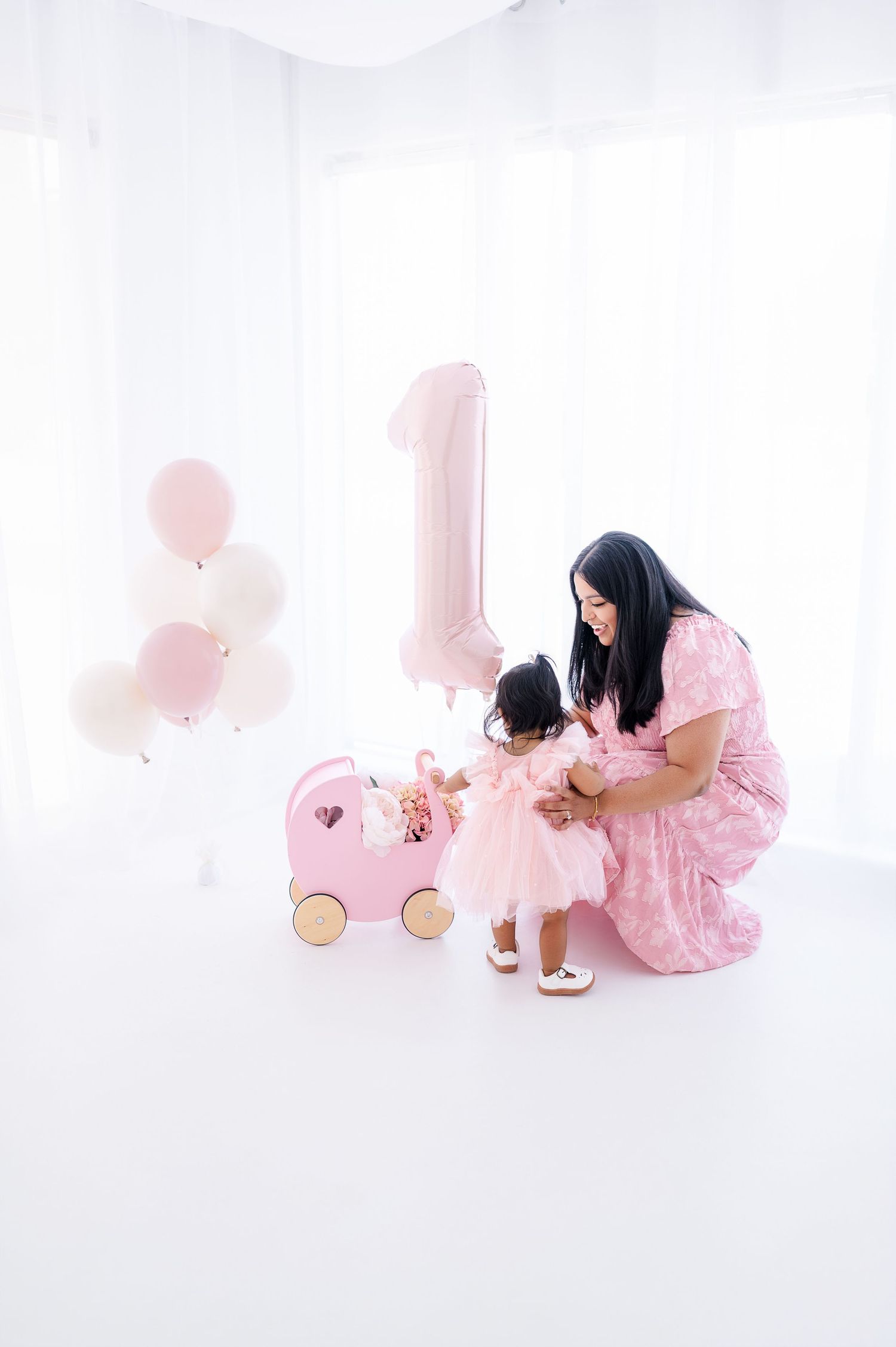 Mother in pink dress kneels beside toddler girl at first birthday party with pink balloons and number one balloon.