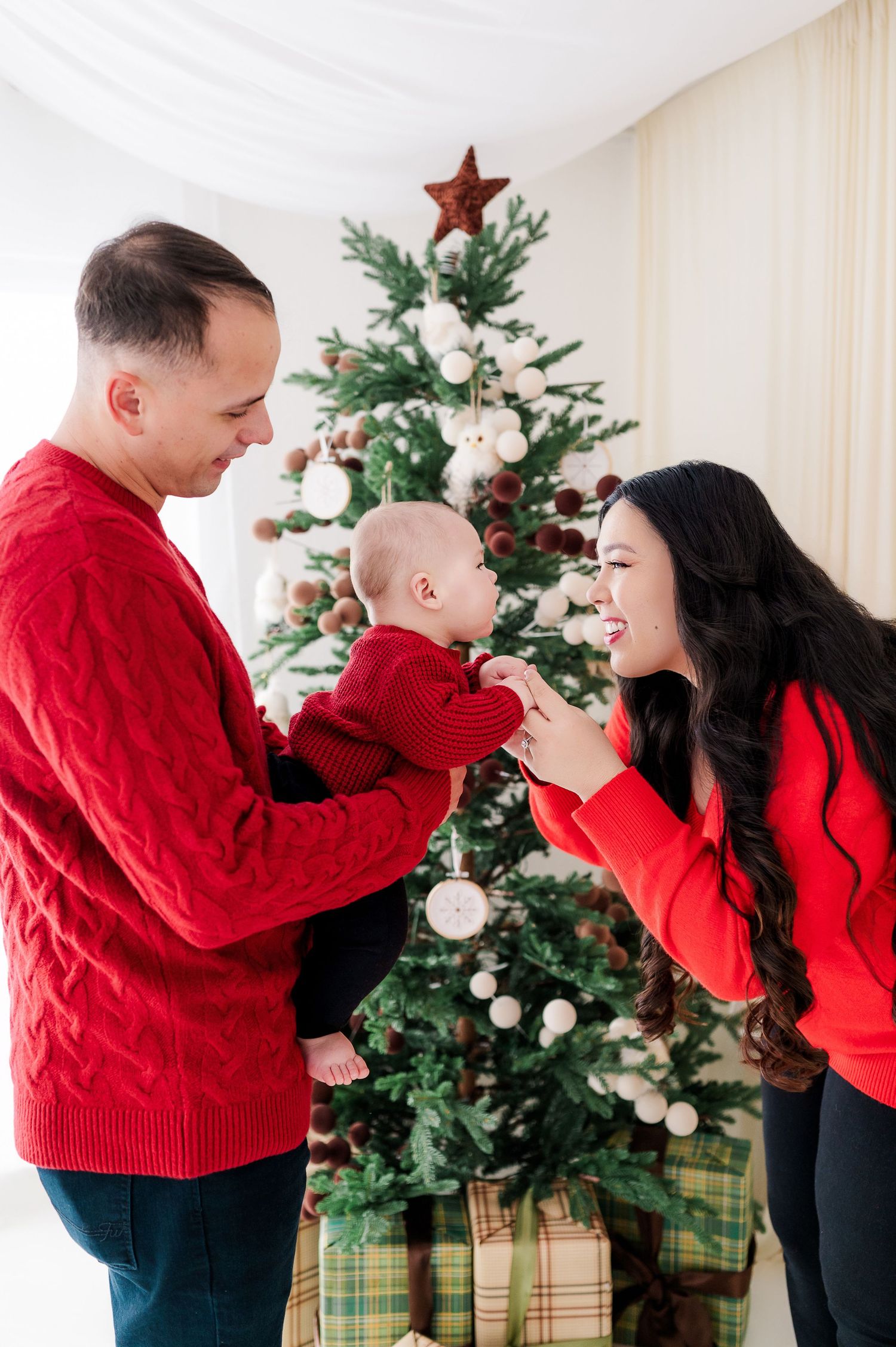 A young family in matching red sweaters celebrates Christmas with their baby in front of a decorated tree with gifts.