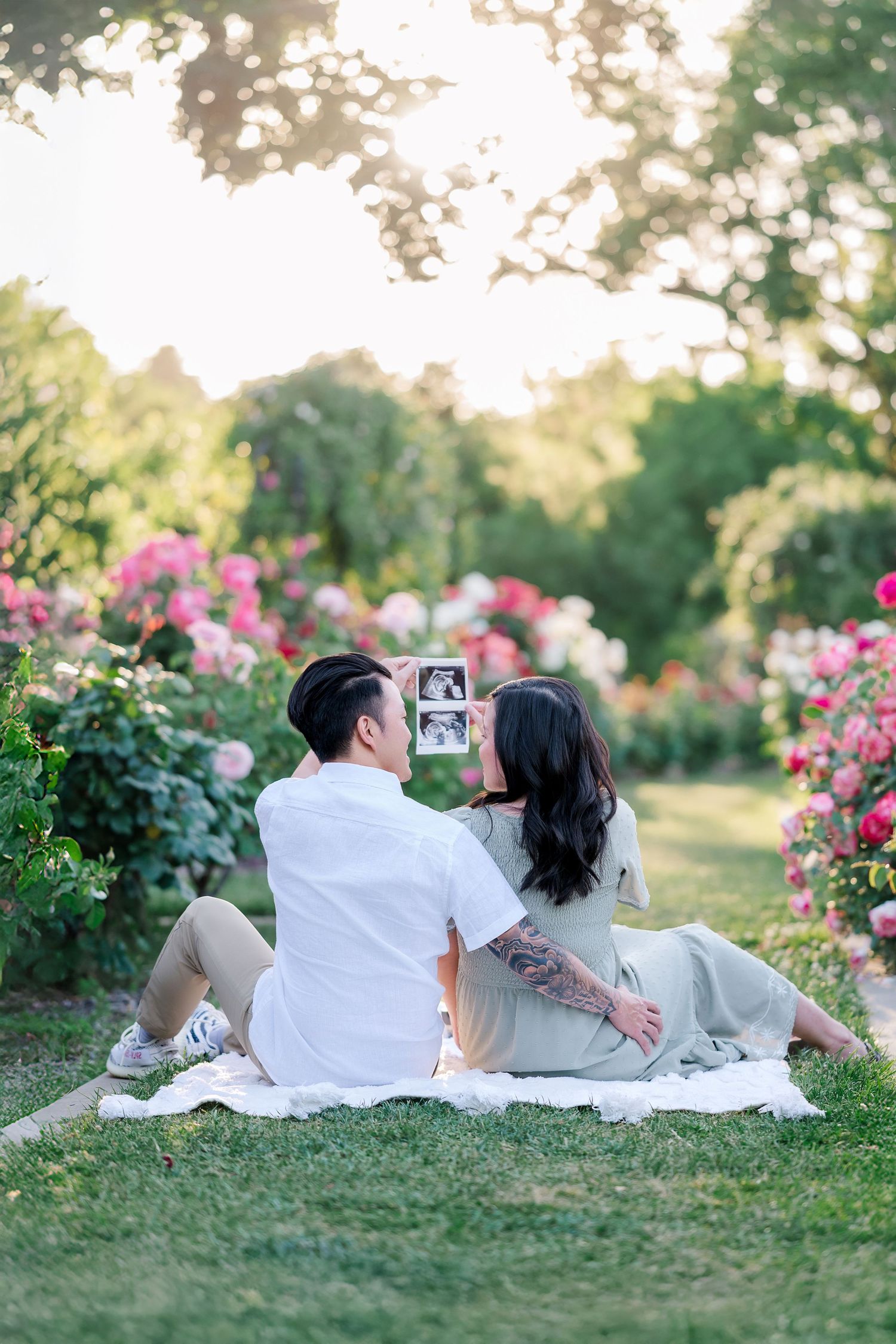 Couple sits on blanket in blooming garden holding ultrasound photo during sunset photoshoot.