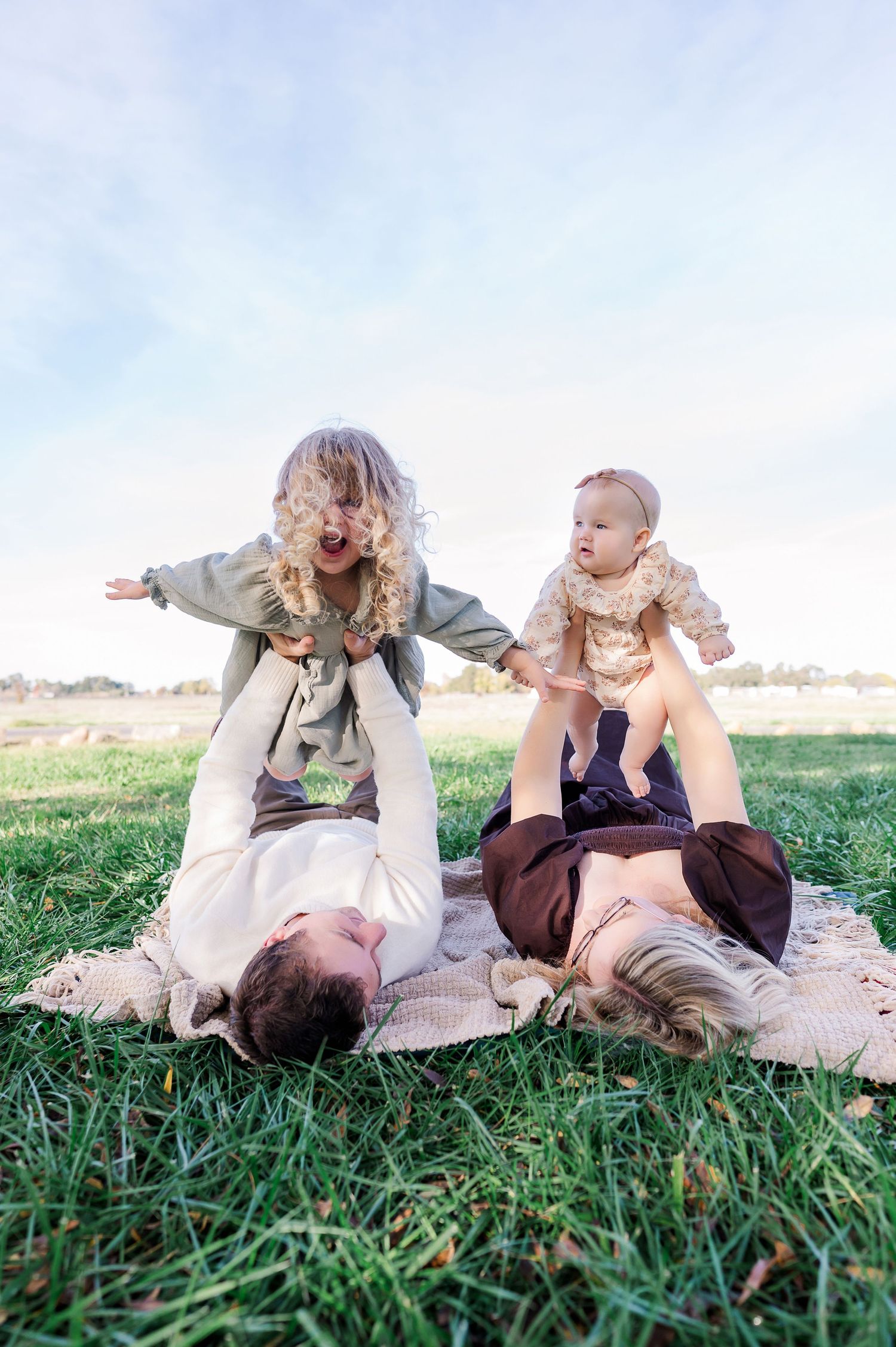Family lying on blanket in grassy field sharing tender moments together during outdoor photoshoot.