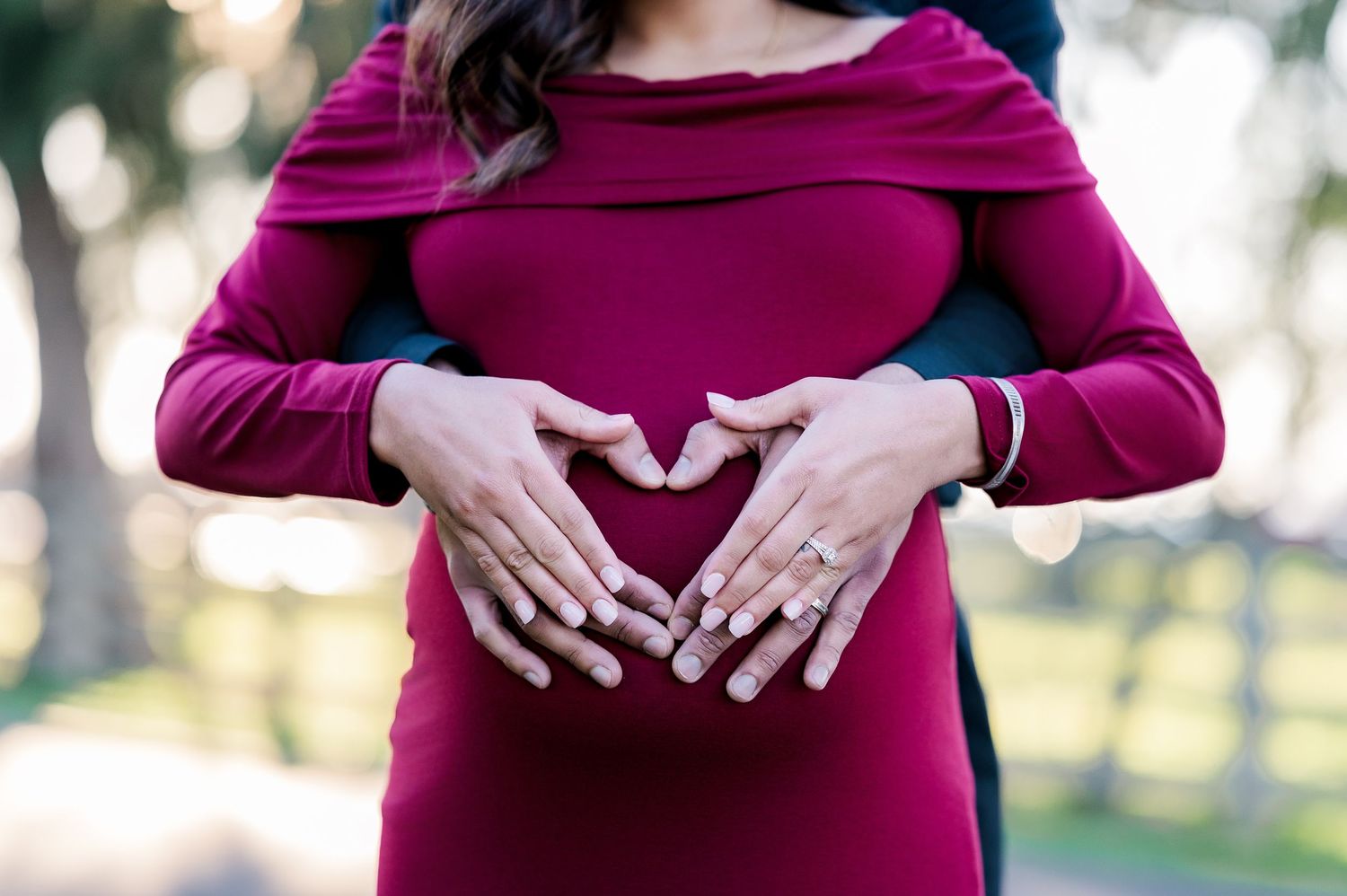 A person in a burgundy dress cradles their pregnant belly while standing outdoors in soft natural lighting.