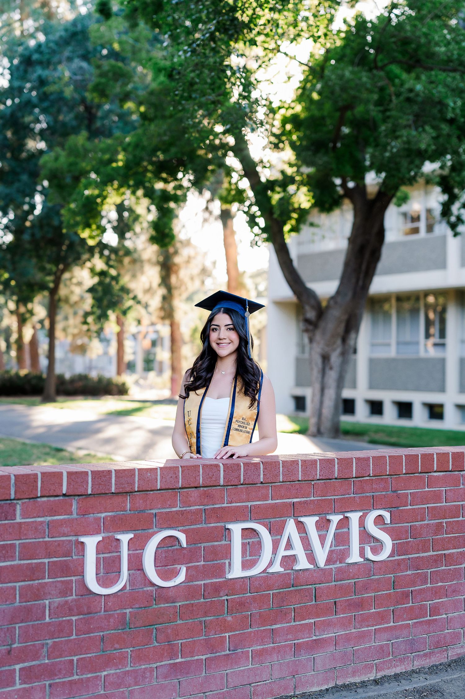 Graduate posing on UC Davis brick entrance sign surrounded by green trees.