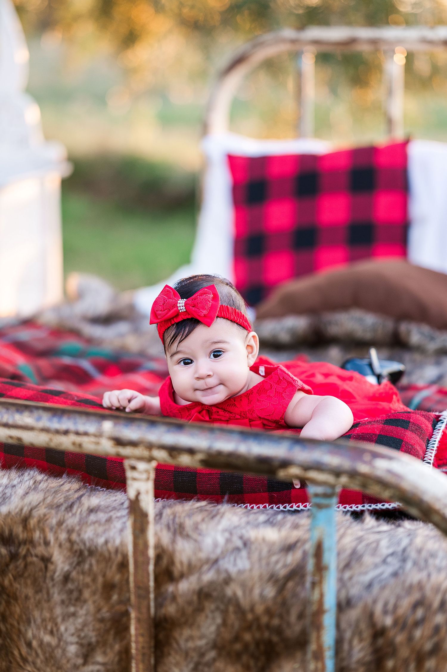 A baby wearing red plaid blanket lies on brown fur backdrop in outdoor holiday photo session.