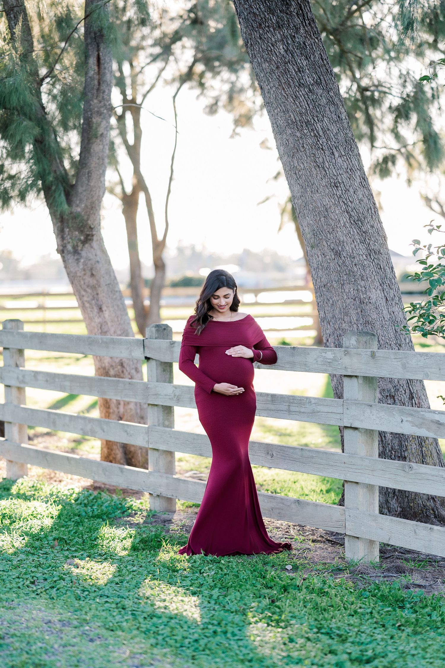 Maternity photography session with burgundy dress against rustic wooden fence and trees.