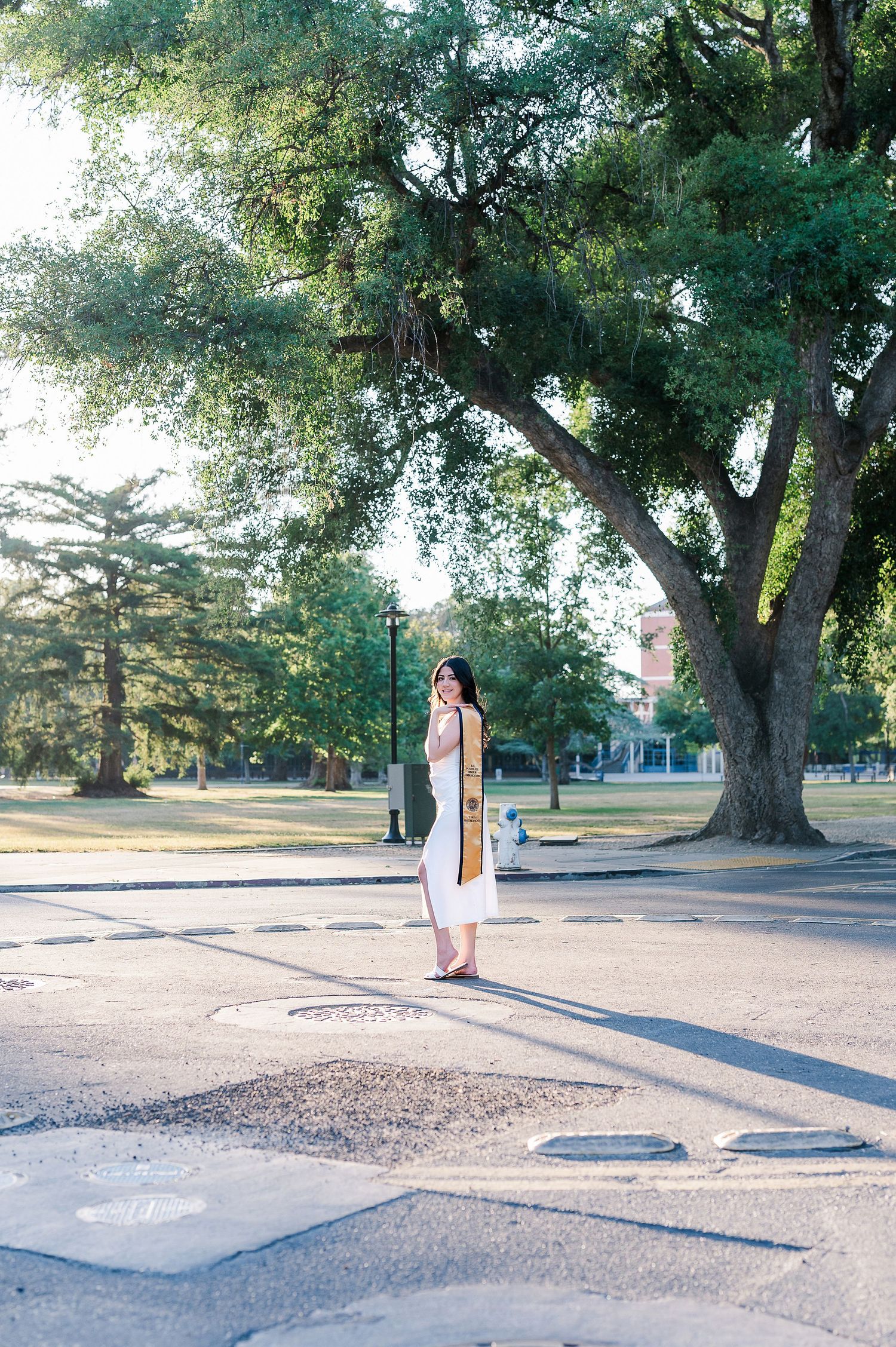 Graduate in white dress walks along a tree-lined pathway during outdoor photo session.