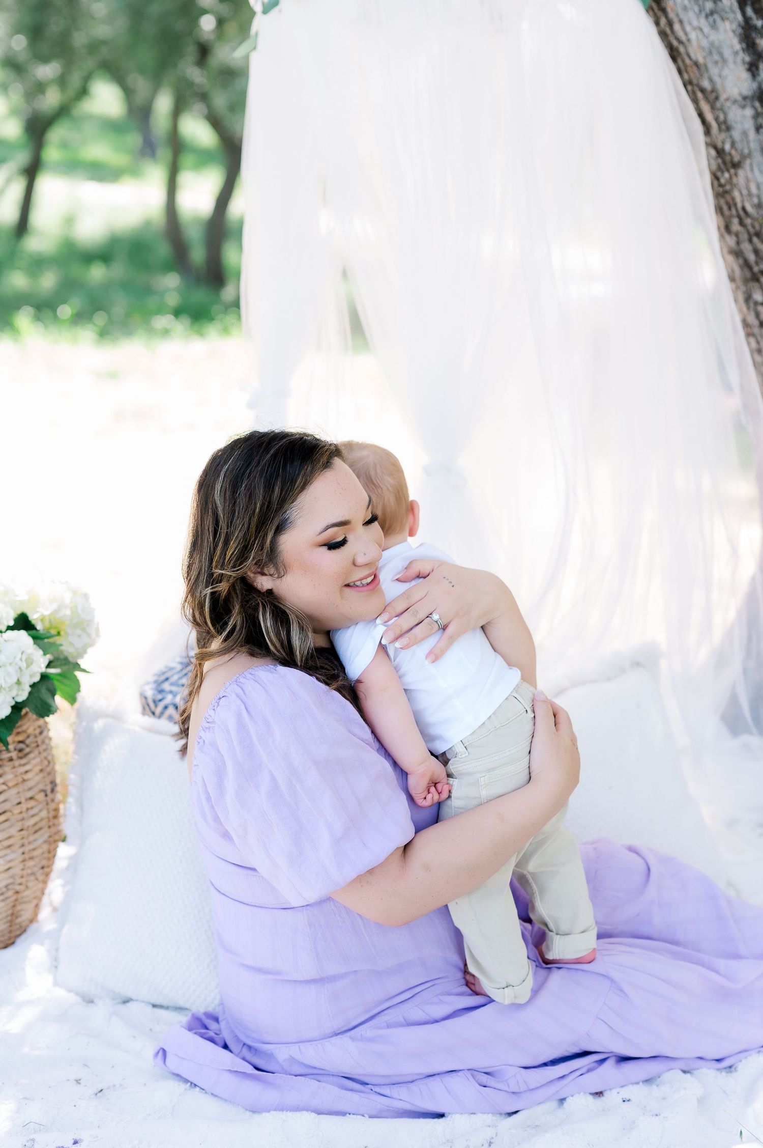 A tender moment captured outdoors as a mother in a lavender dress cuddles with her baby on a white blanket.