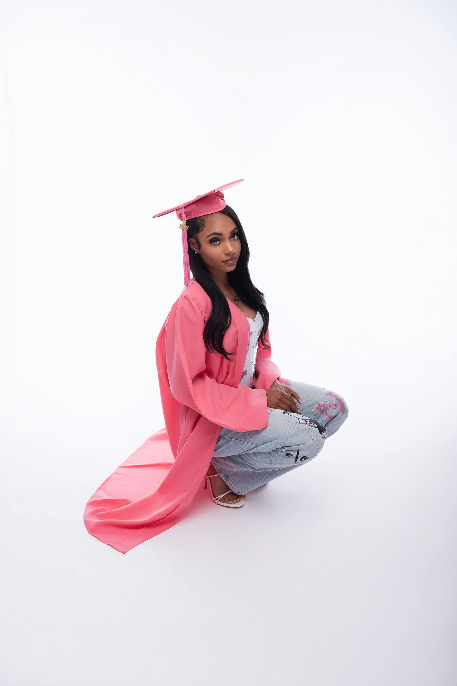 Young woman in pink graduation cap and gown sits cross-legged on white background wearing jeans.