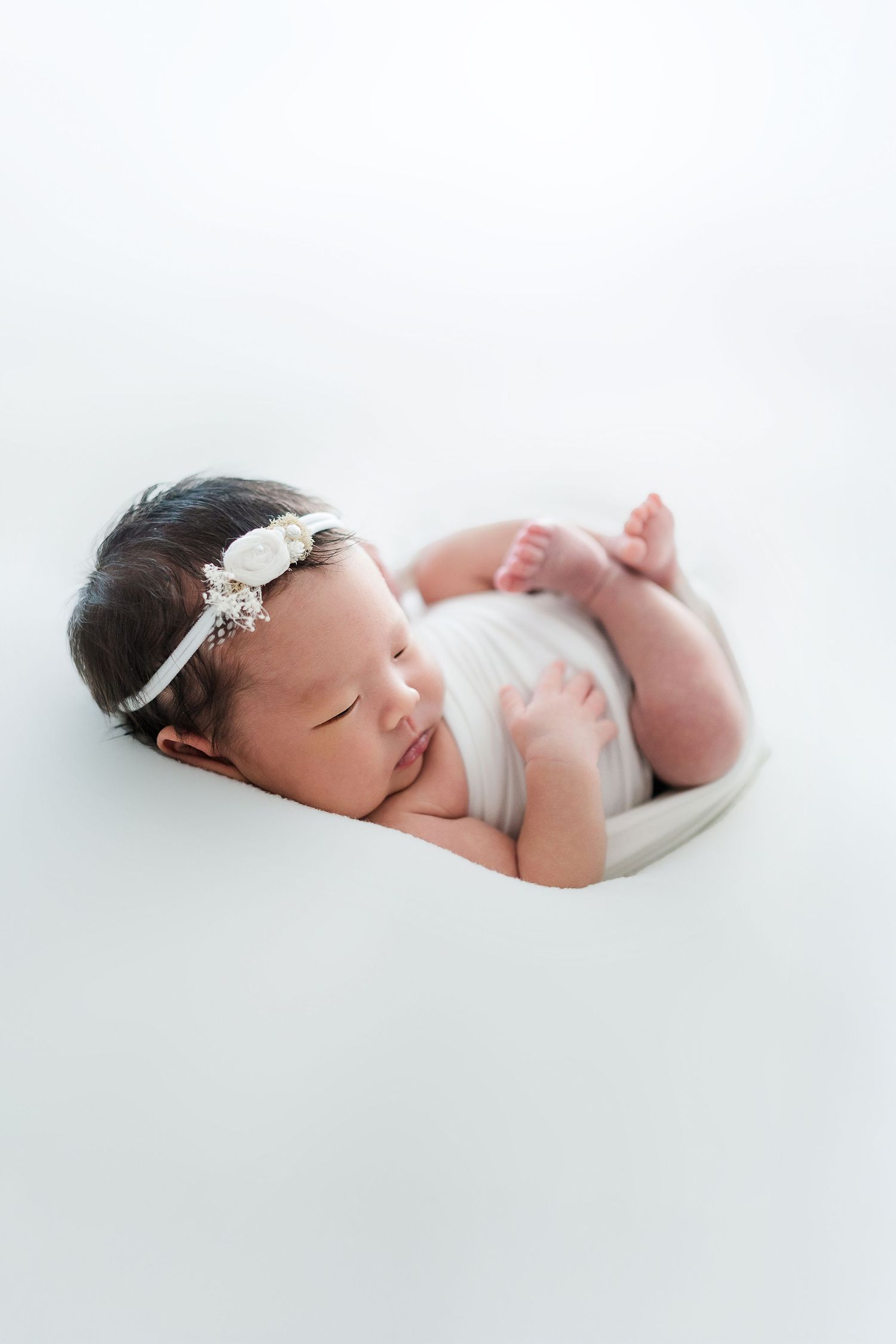Sleeping newborn baby wearing delicate white lace headband resting on white background.