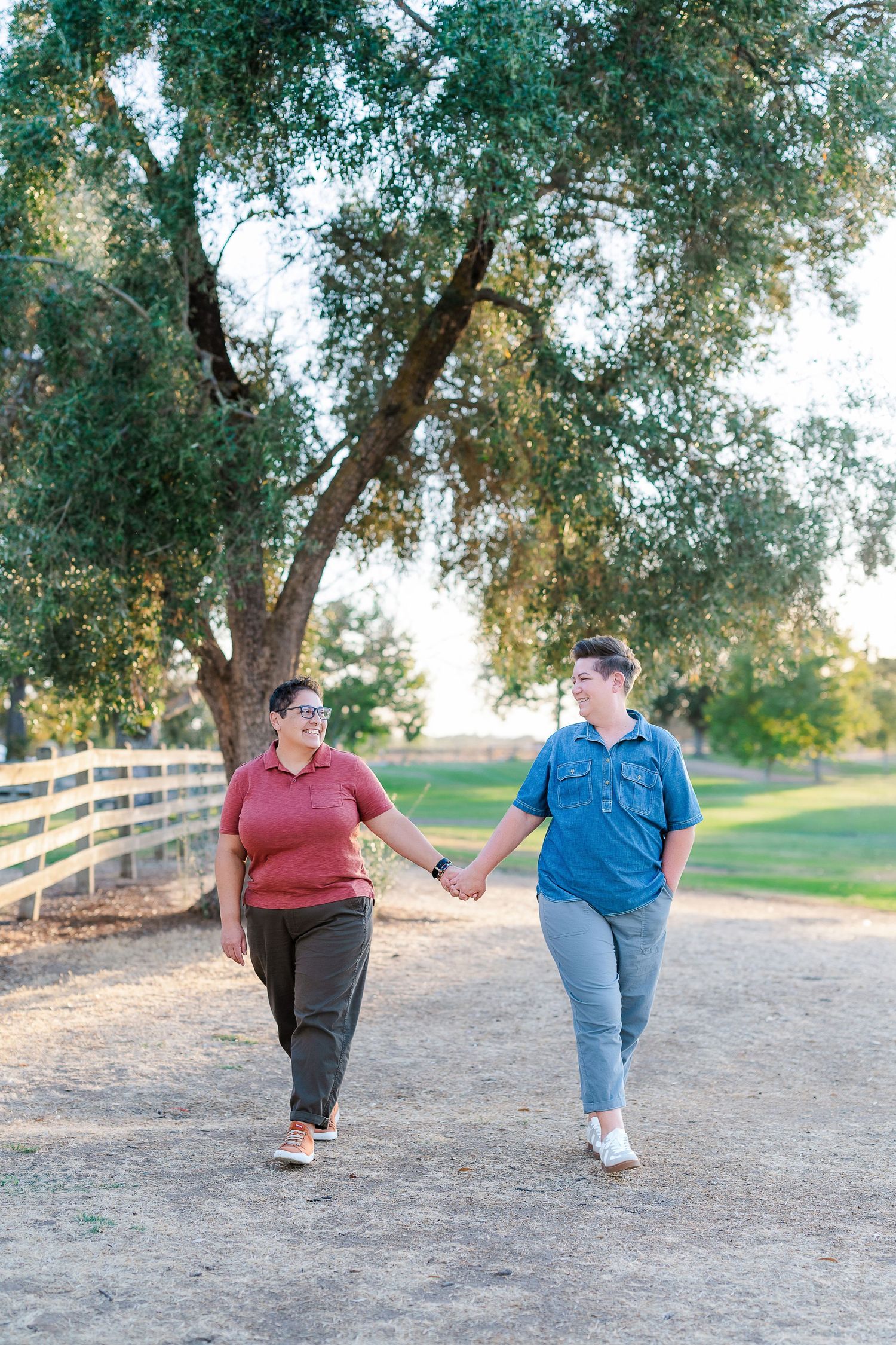 Two people holding hands while walking down dirt path lined with trees and wooden fence.