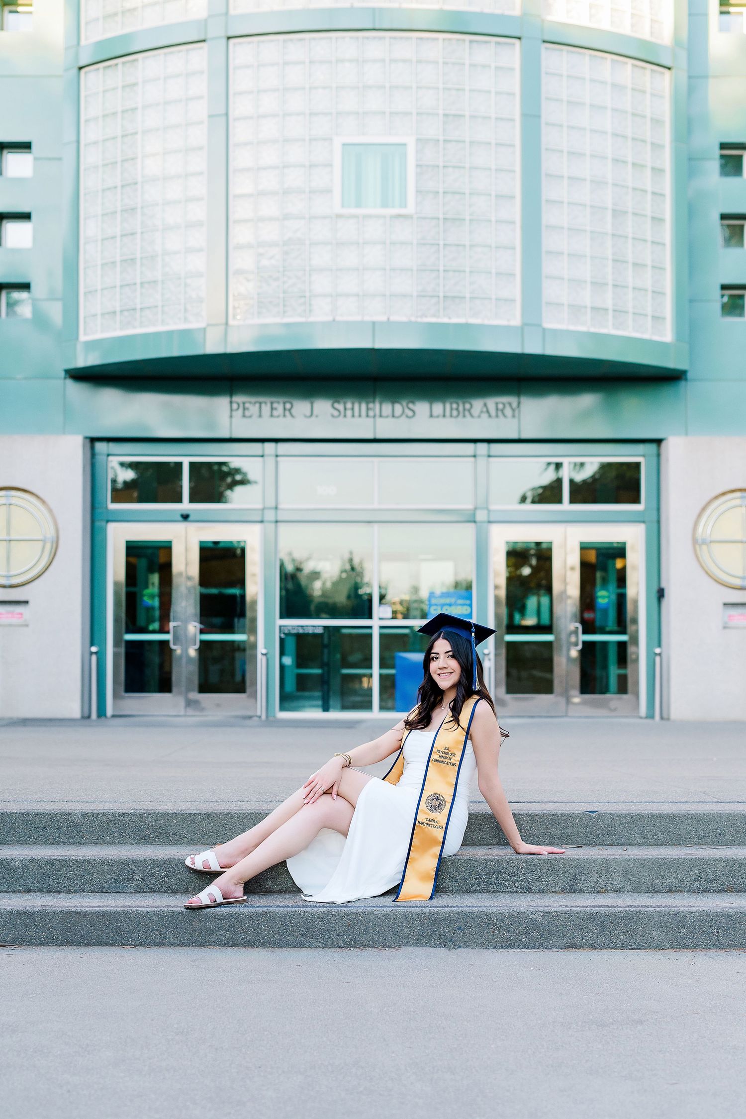 Graduate in white dress and gold sash lounging on steps of a modern campus building.