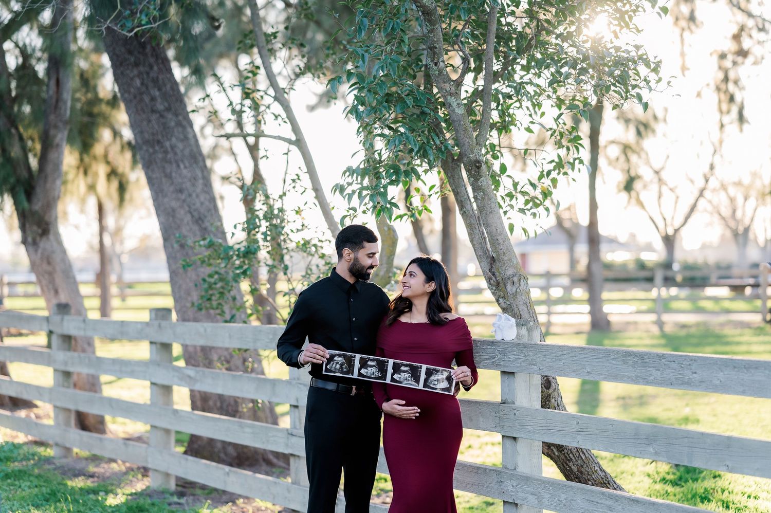 A couple in black and burgundy outfits holds a wooden sign together while standing by a white fence in a park setting.