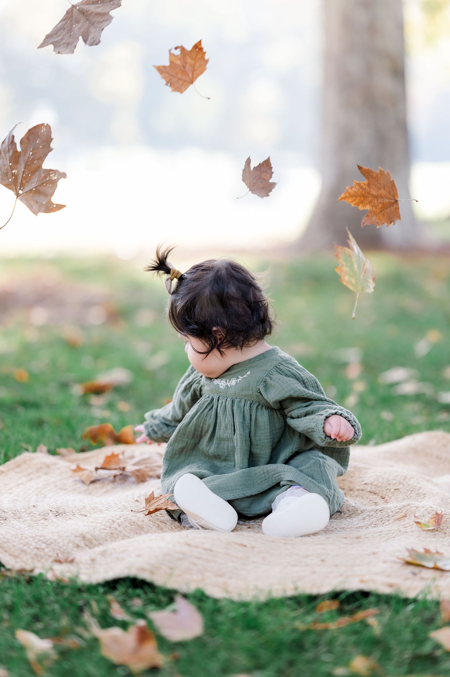 Baby girl in green dress sits on blanket surrounded by falling autumn leaves in a park.