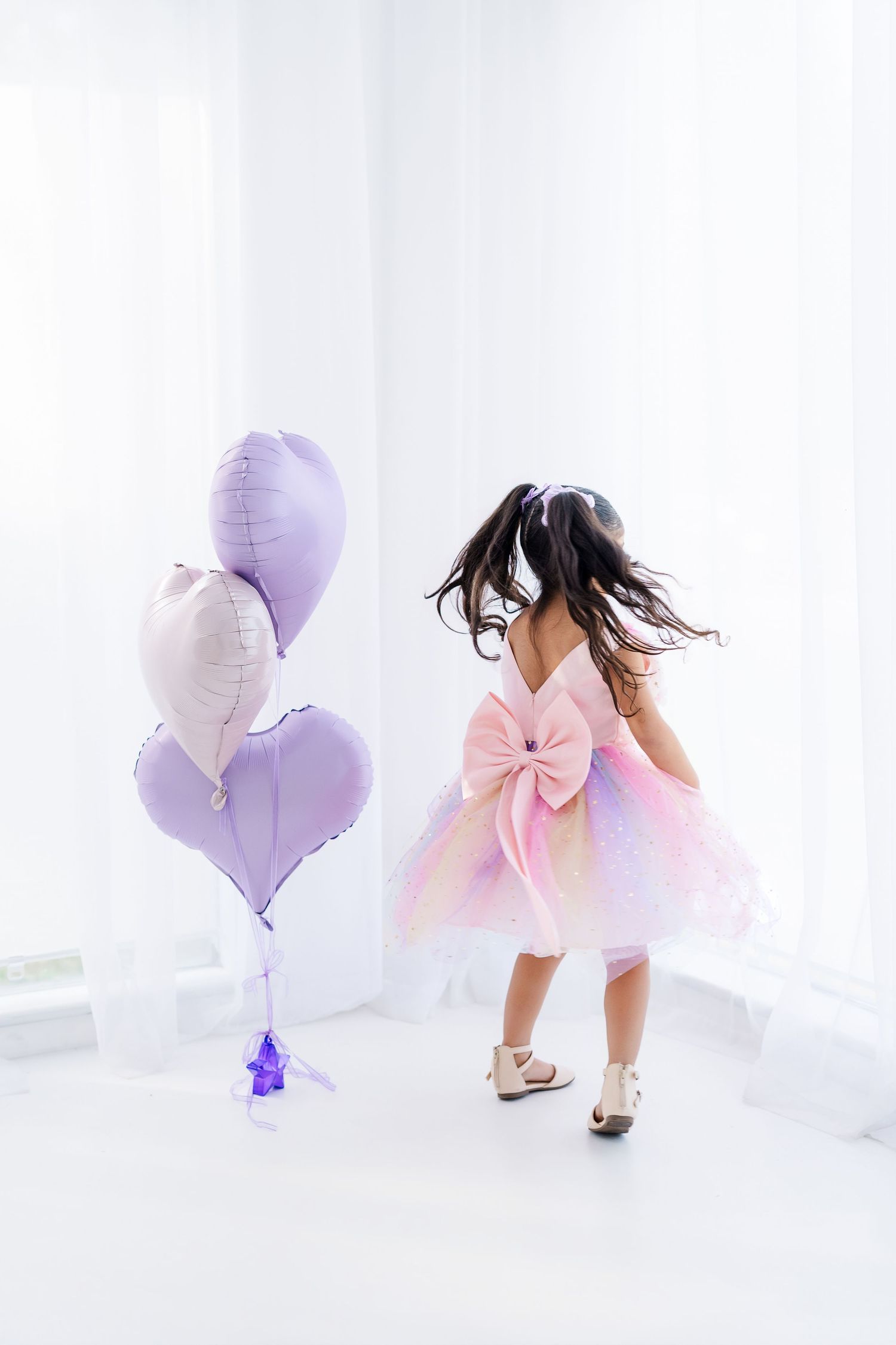 Young girl in colorful rainbow dress with pink bow twirling near purple heart balloons in bright white room.