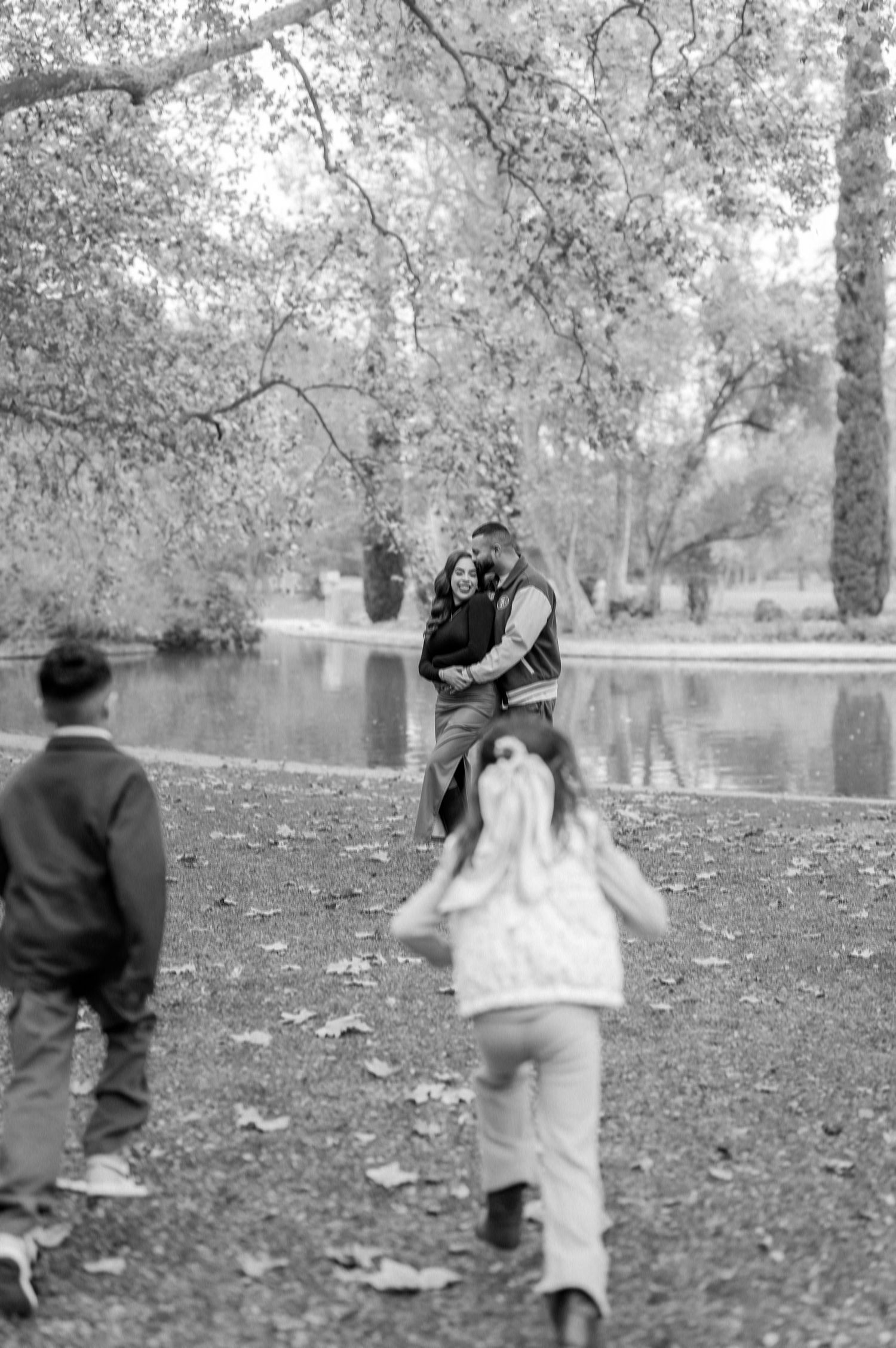 Children playing and running through fallen autumn leaves near a lake in a black and white photograph.