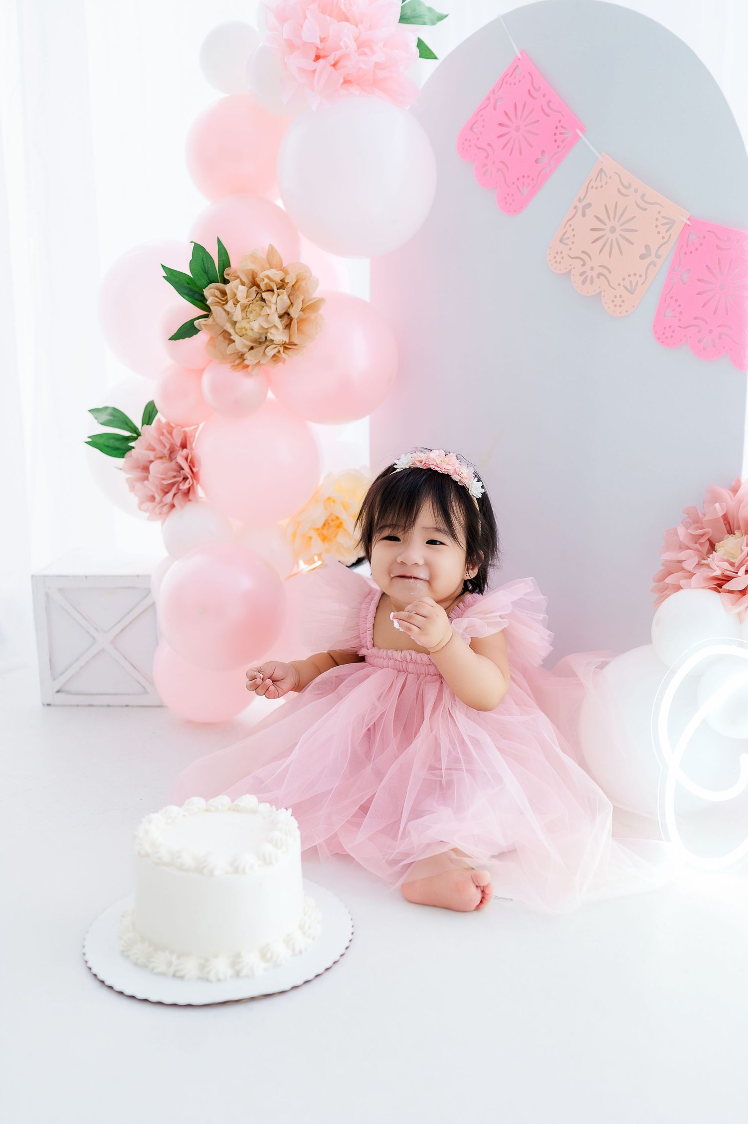 Baby girl in pink tutu dress sits beside white cake at first birthday party with balloon decorations.