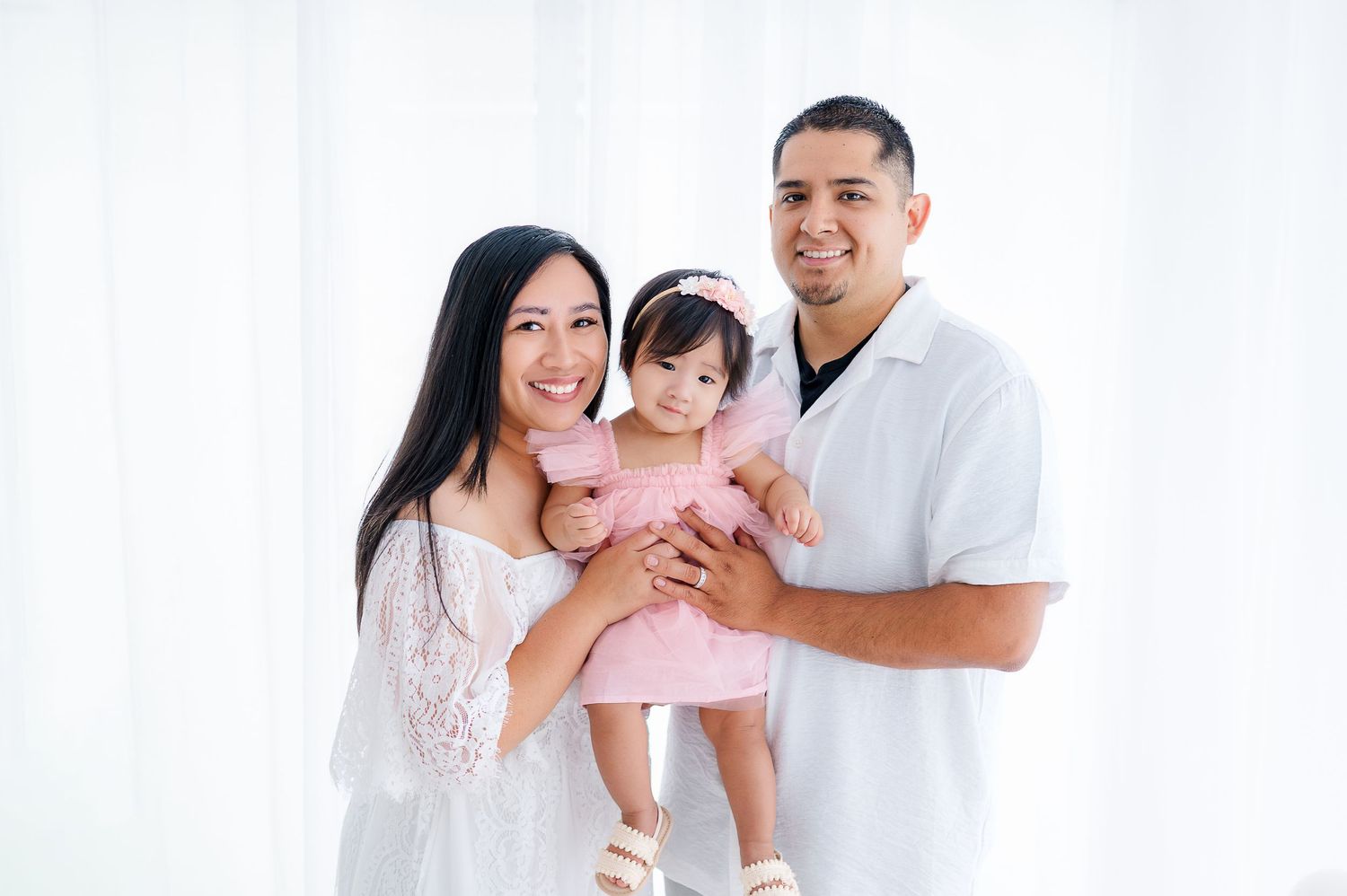 A happy young family with a mother in white, father in white, and toddler girl in pink posing together indoors.
