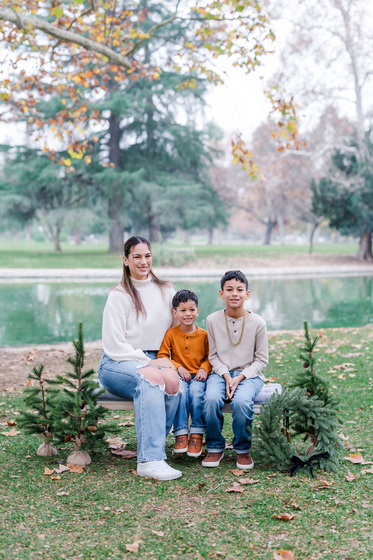 Three siblings sit together on grass near small evergreen trees during a fall photo session outdoors.