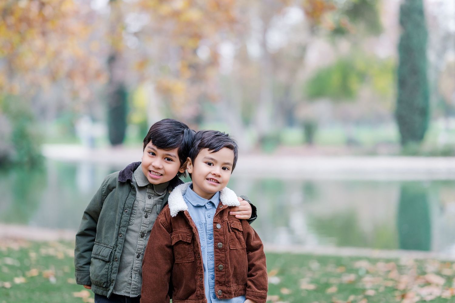 Two people in autumn jackets share a tender moment while walking through a park with fallen leaves.