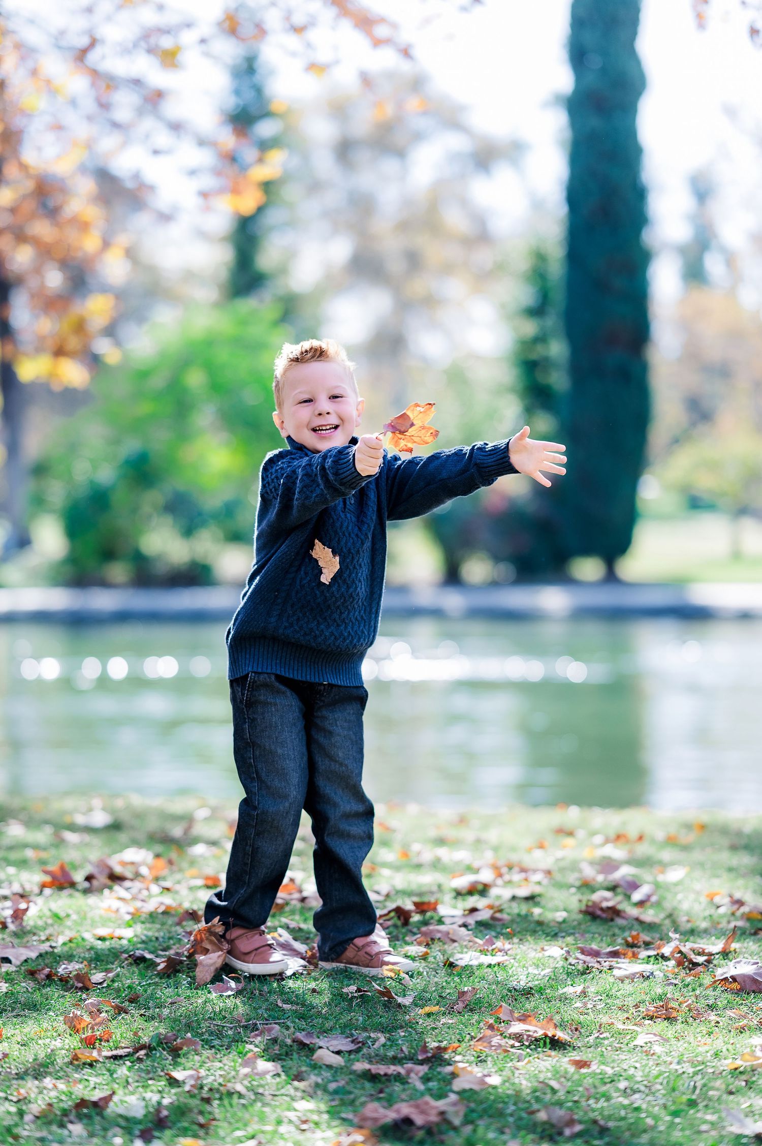 Happy young boy throwing autumn leaves in a park by a pond, wearing a navy sweater and jeans on a fall day.