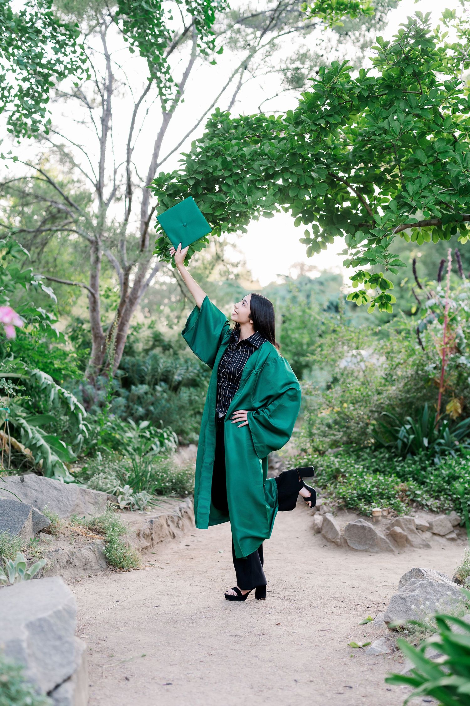 Graduate in emerald green cap and gown celebrates achievement by tossing cap in lush garden setting.