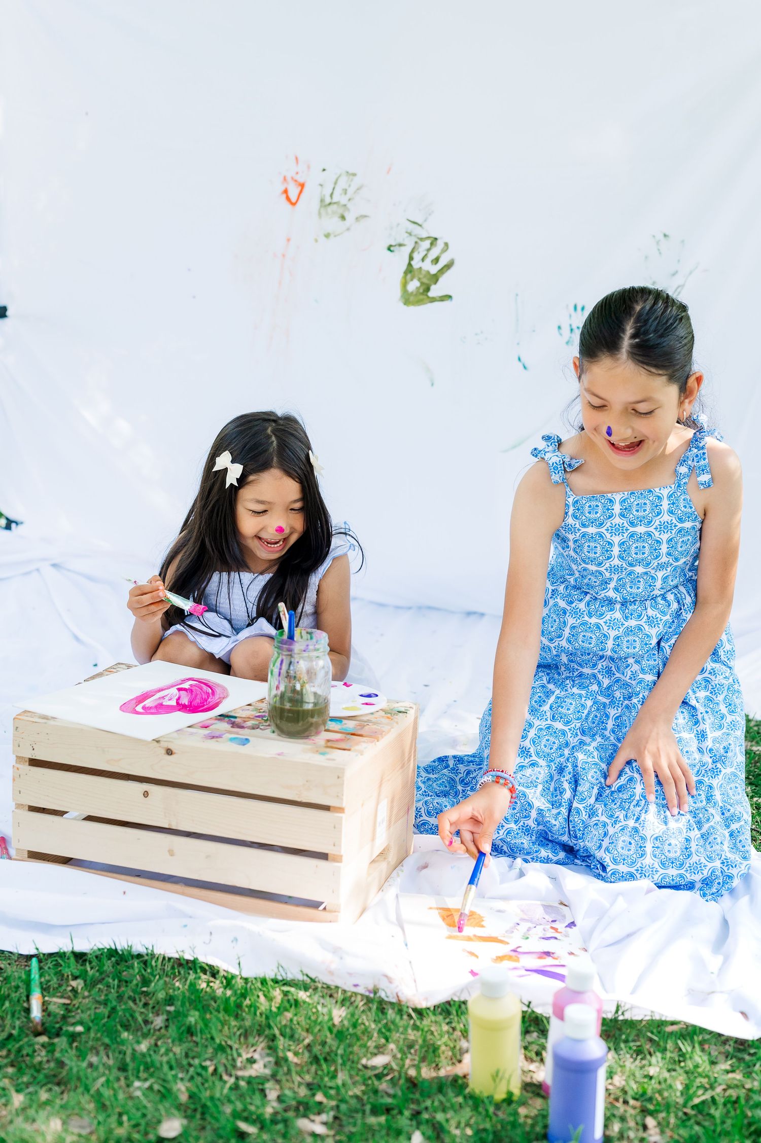 Two children in summer dresses paint together on a white blanket outdoors with colorful art supplies and wooden crates.