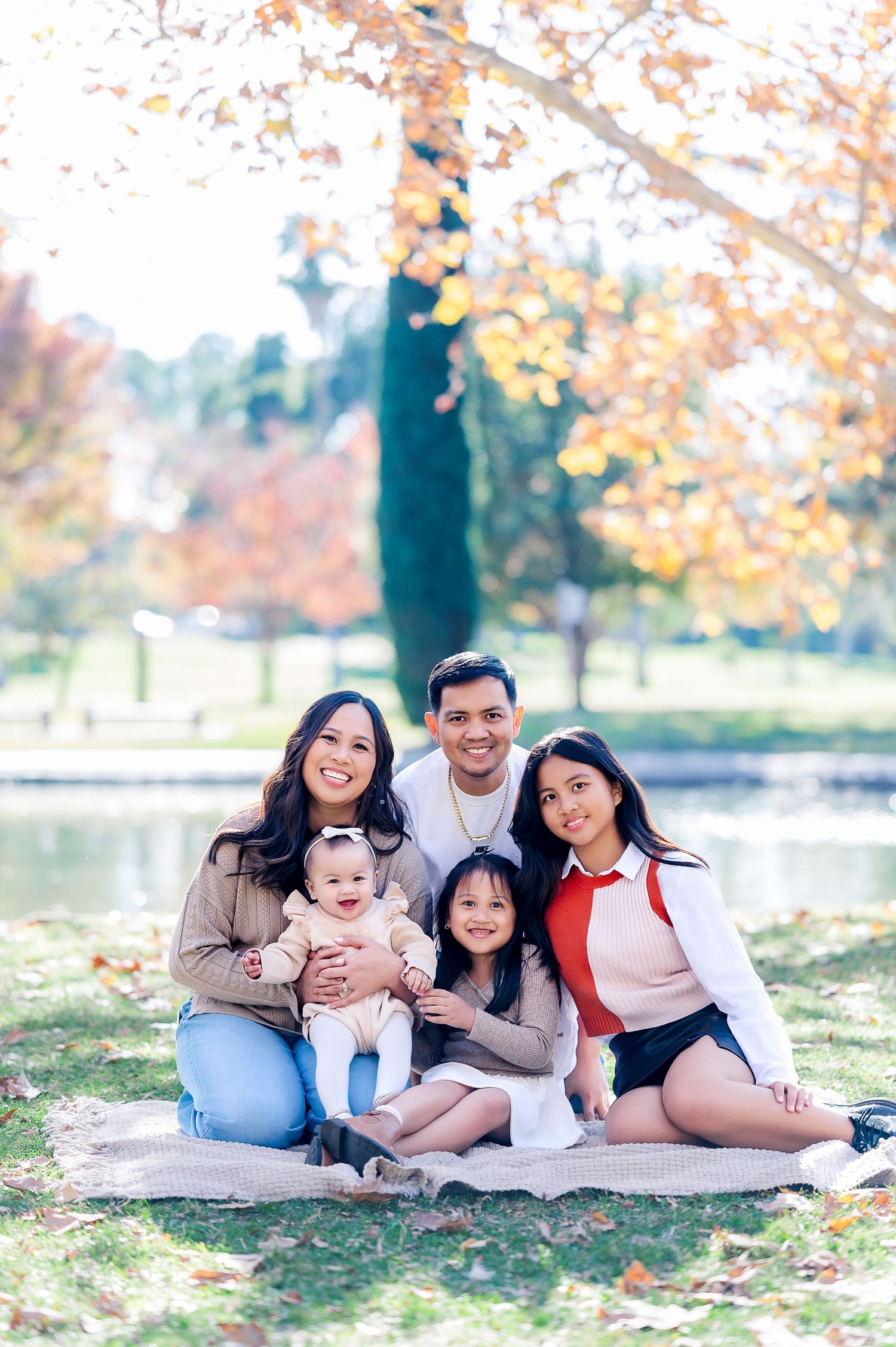 Family sitting on blanket by lake during autumn with colorful fall foliage in background.