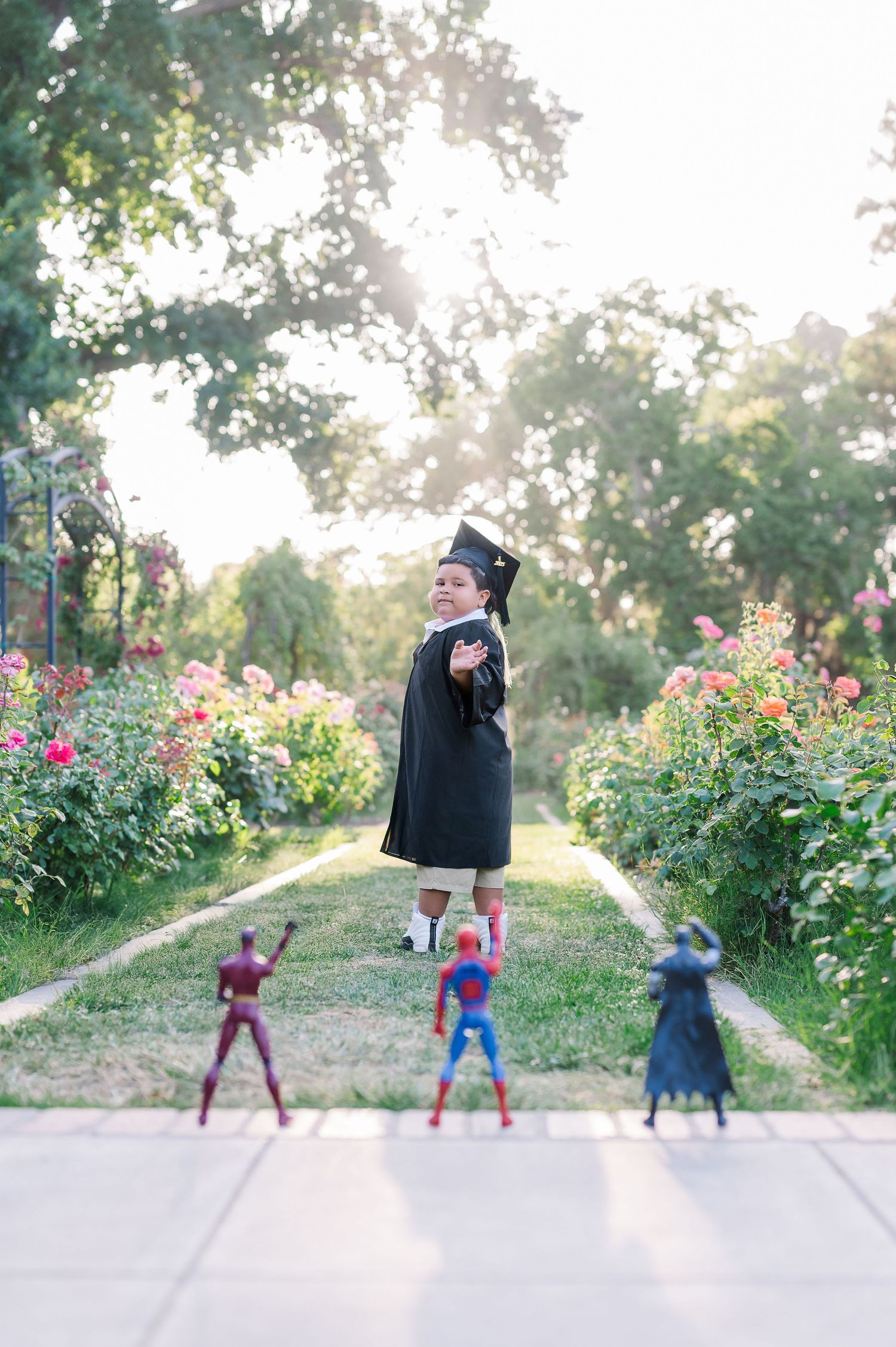 Young child in graduation cap and gown poses outdoors among flowers with superhero toys in foreground.