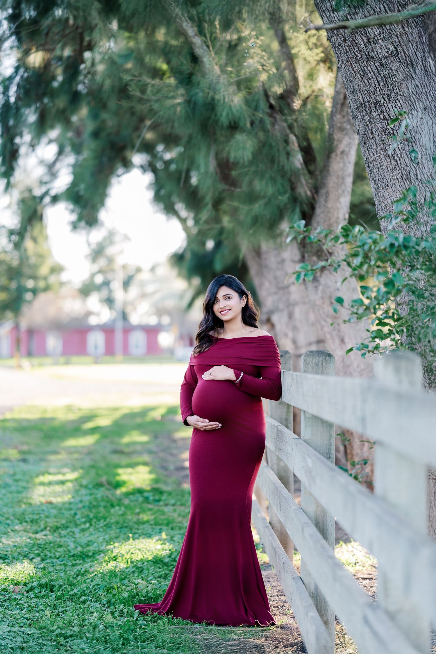 A burgundy dress maternity photo session against a rustic white fence and green trees in a park setting.