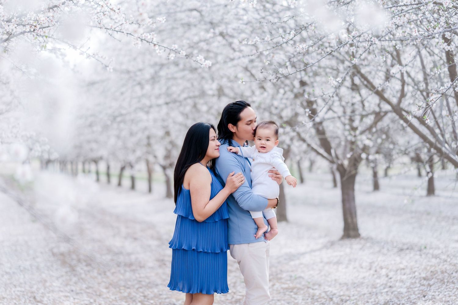 A couple in a blue dress and white shirt embrace in a dreamy orchard setting during winter.